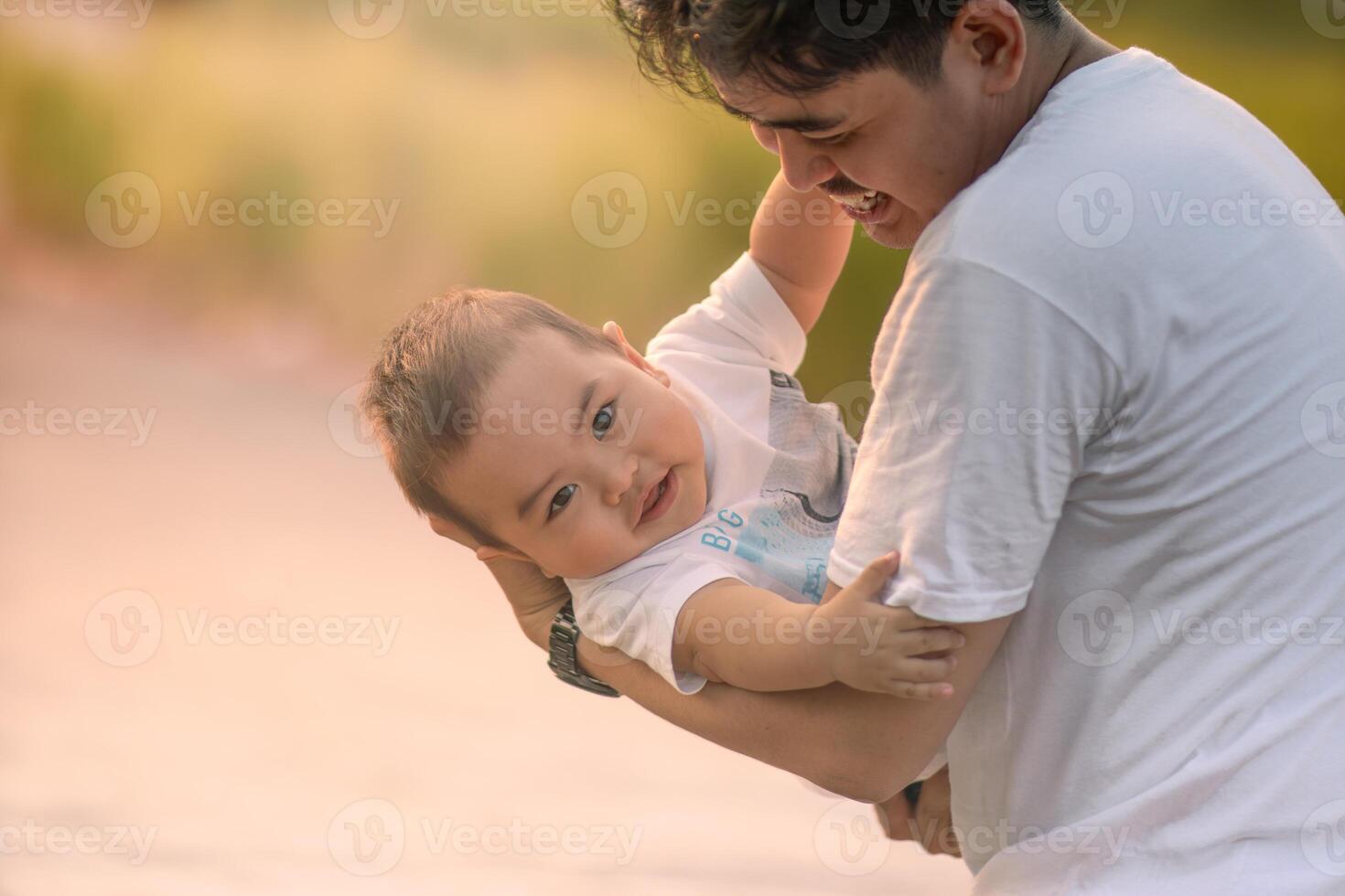 father cradling baby boy and baby boy smiling at camera. father and baby boy wearing white t-shirts. photo with natural background with sunlight in the afternoon.