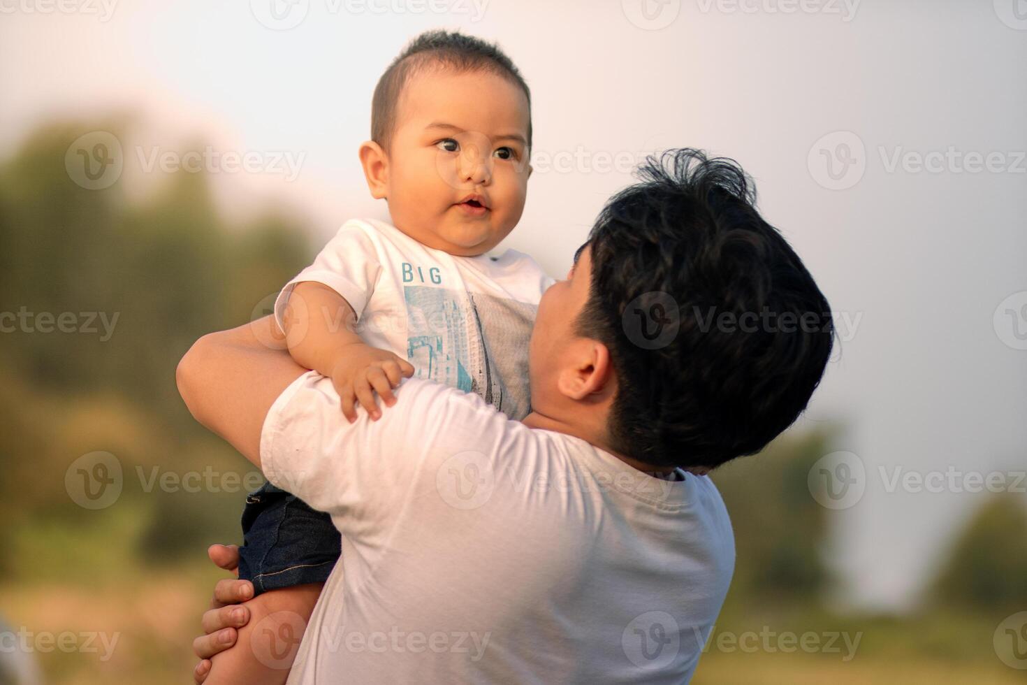 A baby boy is being held by a father and the father looks at the baby boy warmly with a smile. father holding baby boy outdoors in open yard with his back facing the camera. photo