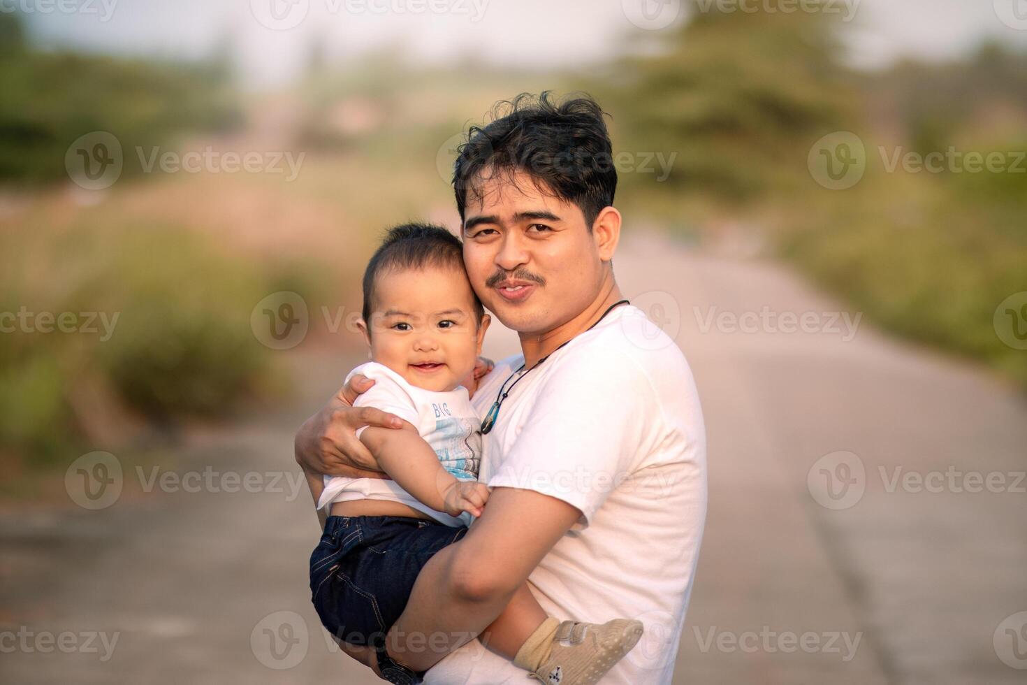 father holding and hugging baby boy outside the house. they smile facing the camera. background of natural atmosphere in the afternoon. photo