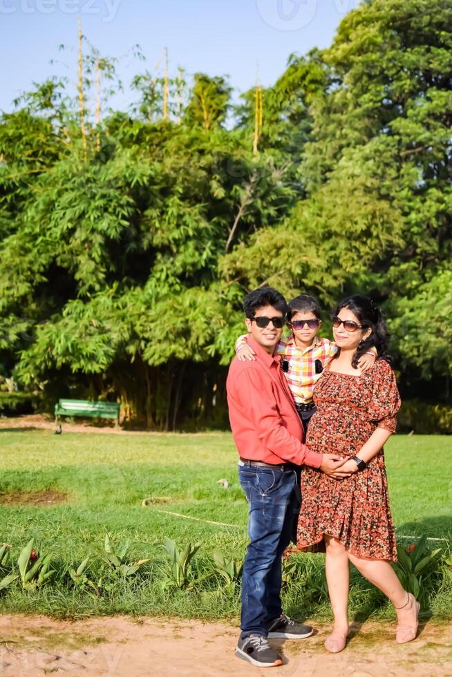 Indian couple posing for maternity baby shoot with their 5 year old kid. The couple is posing in a lawn with green grass and the woman is flaunting her baby bump in Lodhi Garden in New Delhi, India photo