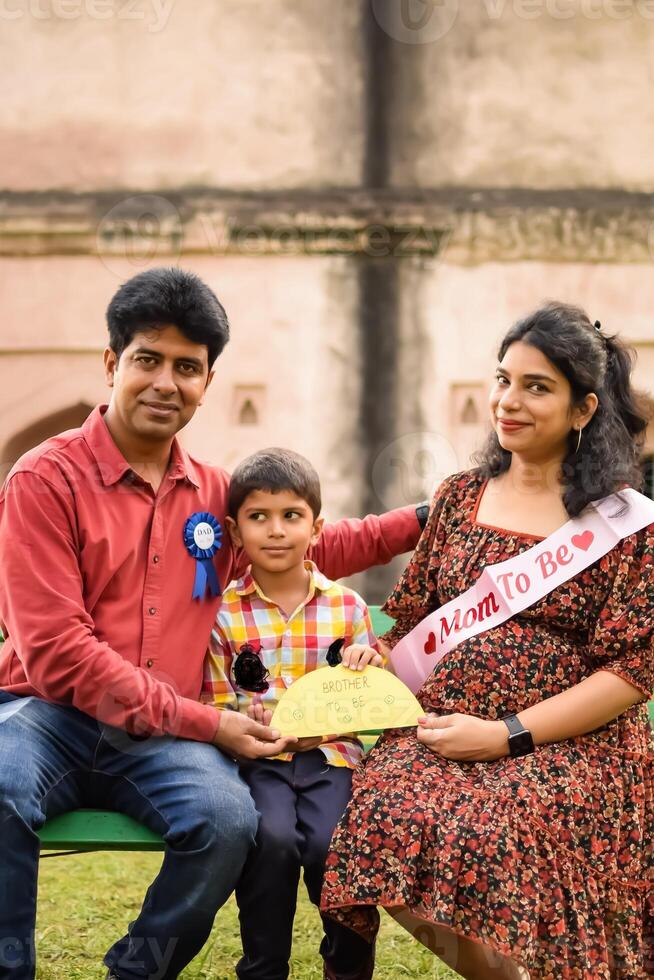 Indian couple posing for maternity baby shoot with their 5 year old kid. The couple is posing in a lawn with green grass and the woman is flaunting her baby bump in Lodhi Garden in New Delhi, India photo