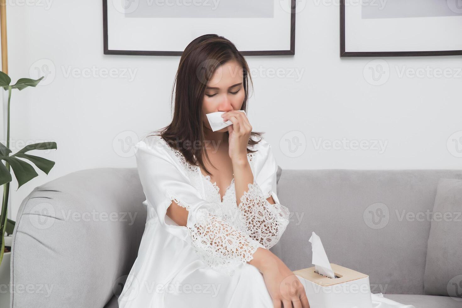 An Asian woman, adorned in a long nightgown and a white satin robe, uses a tissue to cover her nose due to a runny nose caused by nighttime allergies from the changing weather. photo