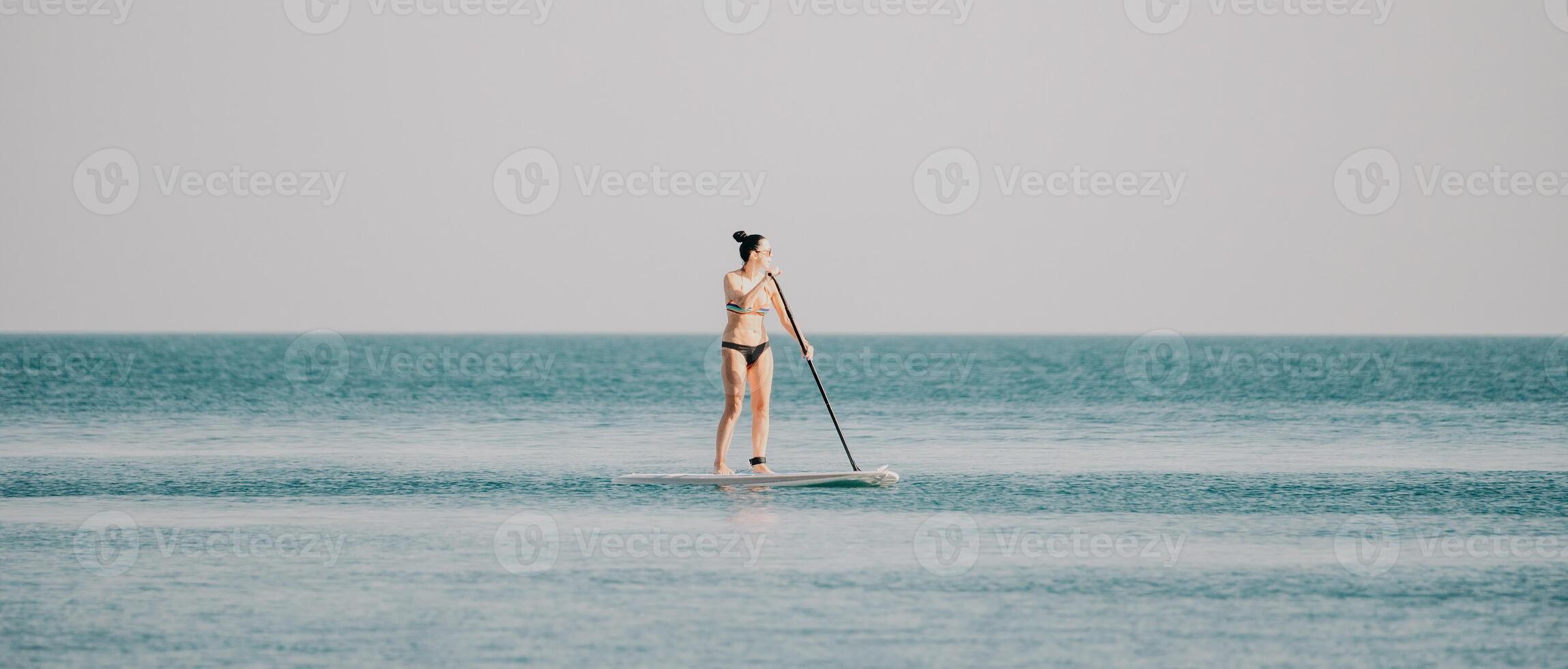 Sea woman sup. Silhouette of happy middle aged tanned woman in rainbow bikini, surfing on SUP board, confident paddling through water surface. Idyllic sunset. Active lifestyle at sea or river. photo
