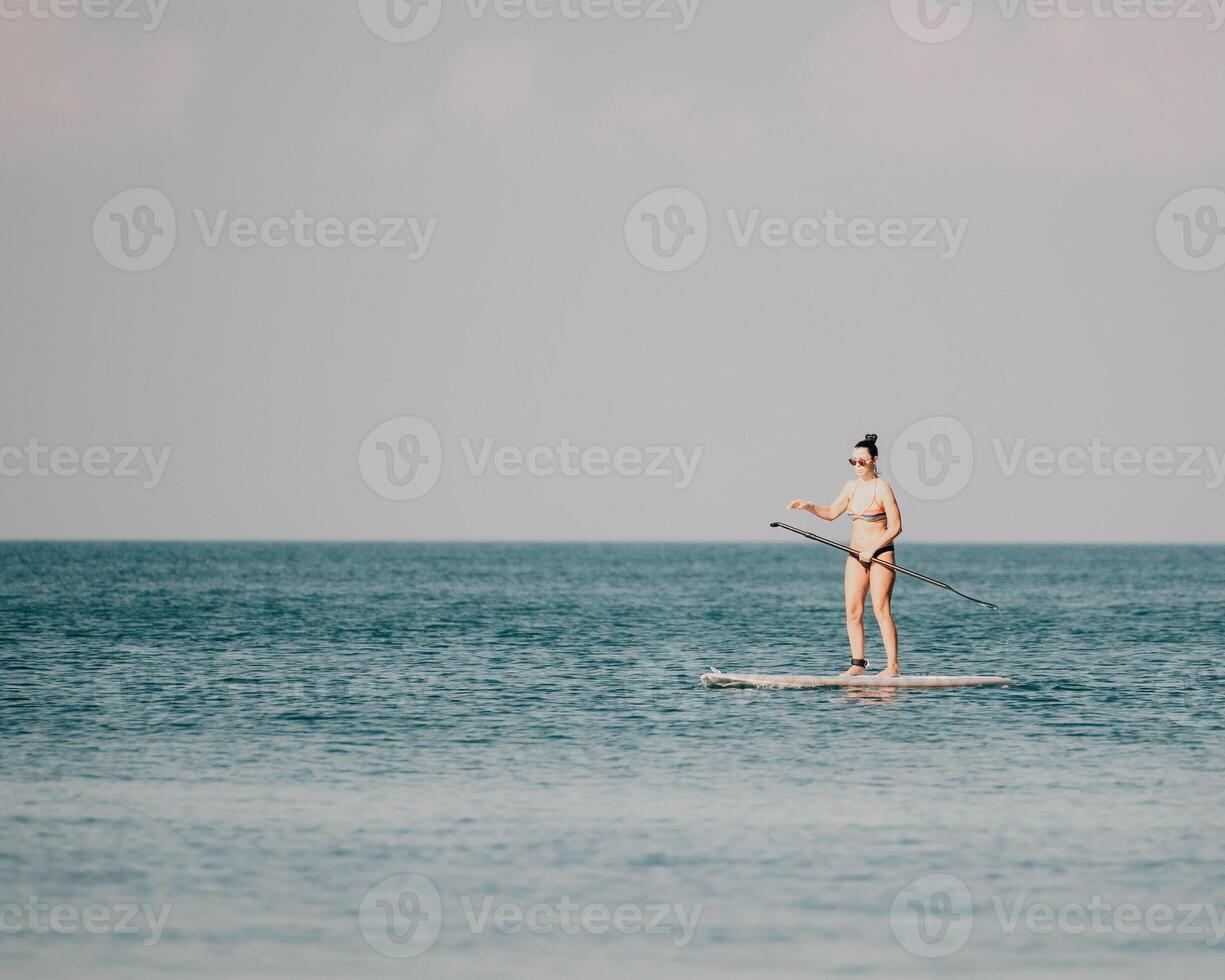 Sea woman sup. Silhouette of happy middle aged tanned woman in rainbow bikini, surfing on SUP board, confident paddling through water surface. Idyllic sunset. Active lifestyle at sea or river. photo