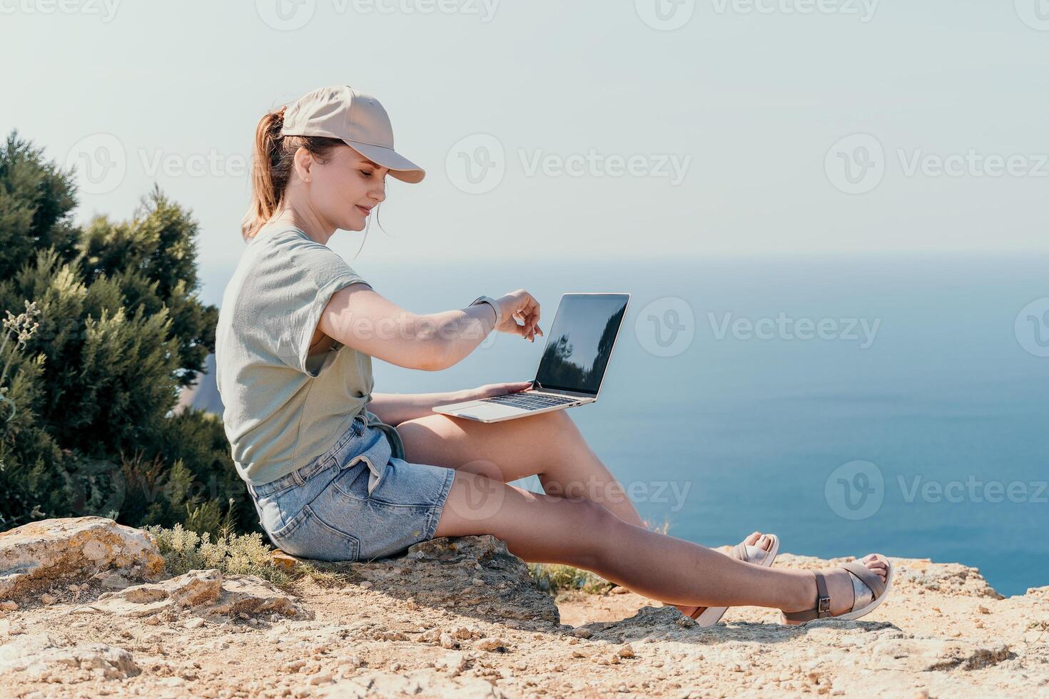 Young Woman Working on Laptop While Sitting on a Cliff Overlooking the Ocean photo