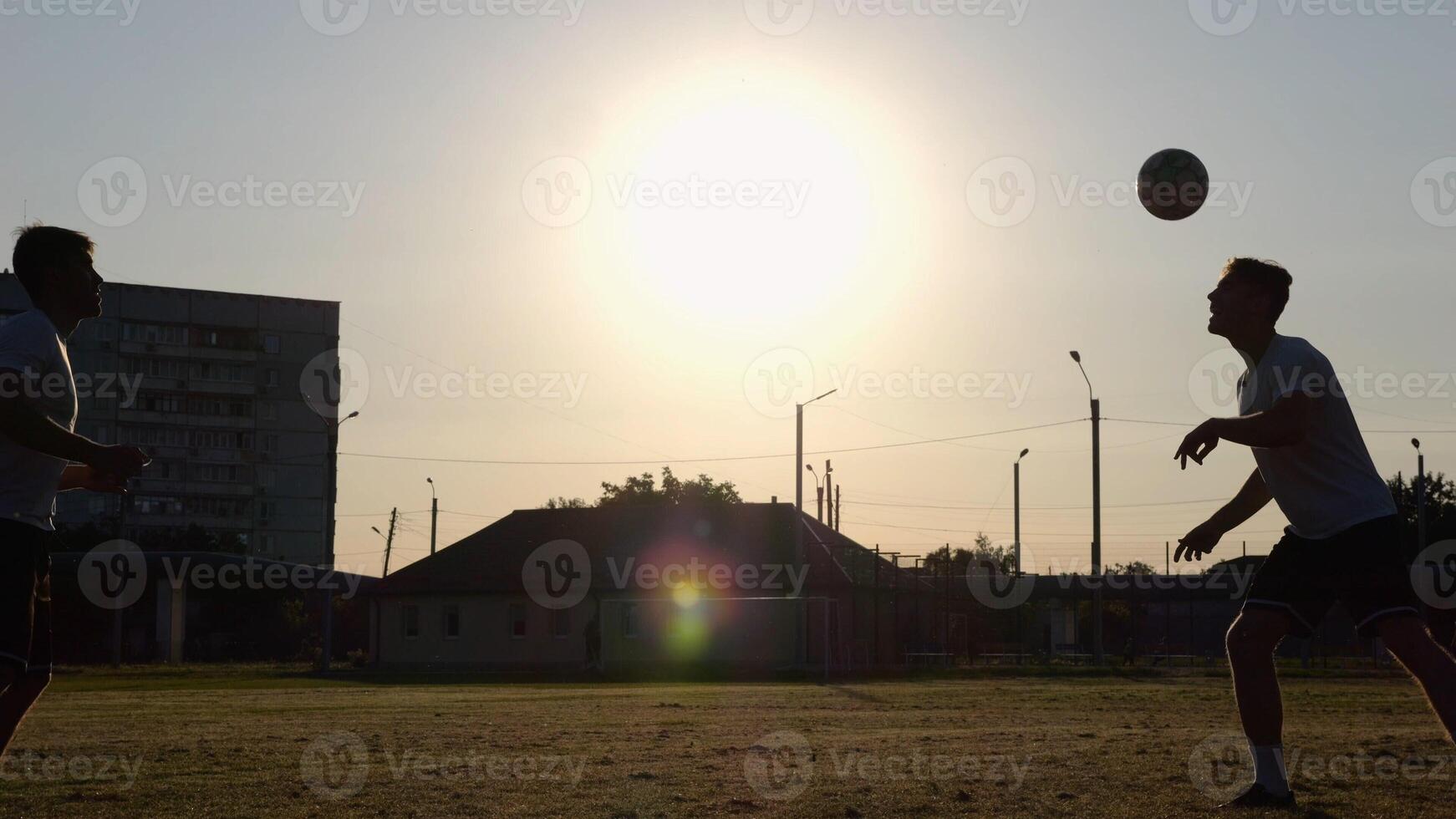 Silhouette of professional footballers kicking soccer ball with their heads on stadium at sunset. Two sportsmen showing tricks with ball while passing it to each other at field. Freestyle football photo