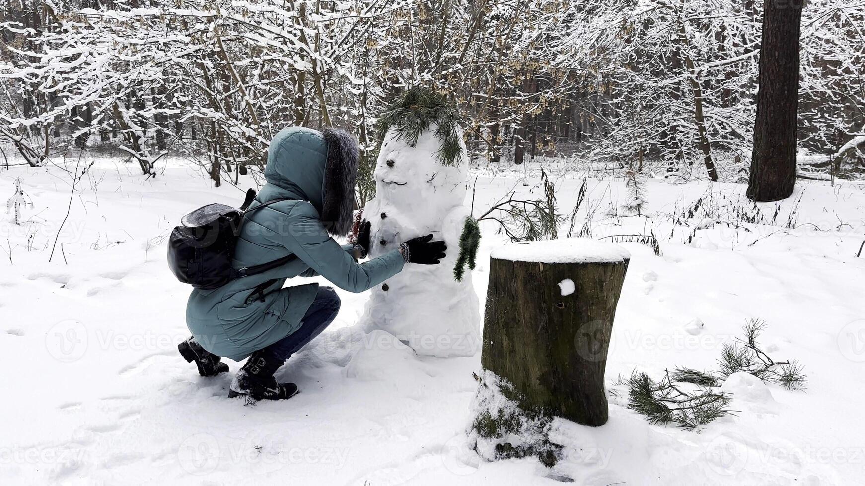 Happy woman building snowman at winter forest. Young girl having active leisure at snowy park. Cheerful female having fun outdoor. Concept of a winter vacation. photo