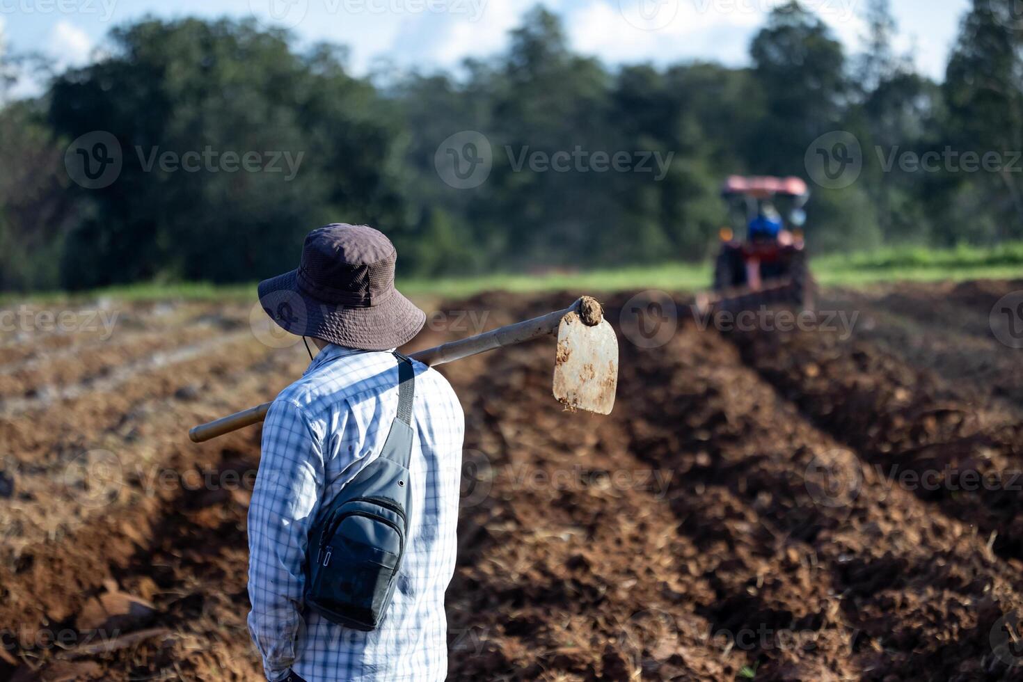 Asian farmer is carrying hoe to plough tilled soil ready for planting in the countryside along with tractor during summer for potato plantation and agriculture photo