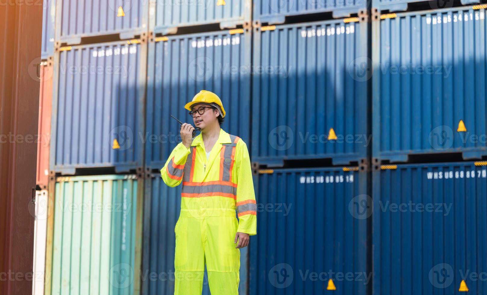Asian man worker talking on walkie talkie for control loading containers box while standing in front of blue containers at cargo shipping yard photo