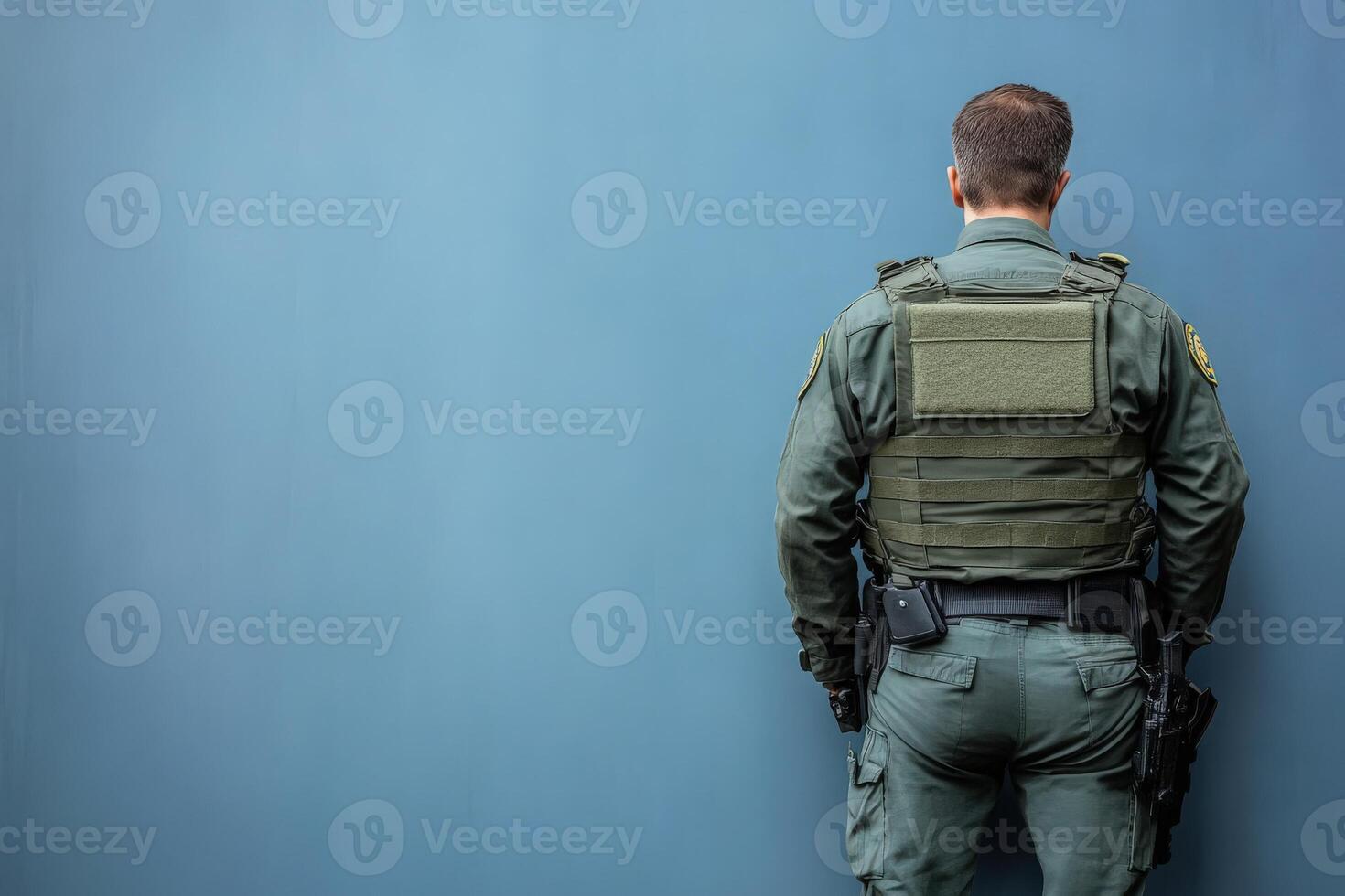 A border control officer is positioned with their back to viewers, wearing a tactical uniform. The clean blue background emphasizes their role and readiness during a duty shift. photo