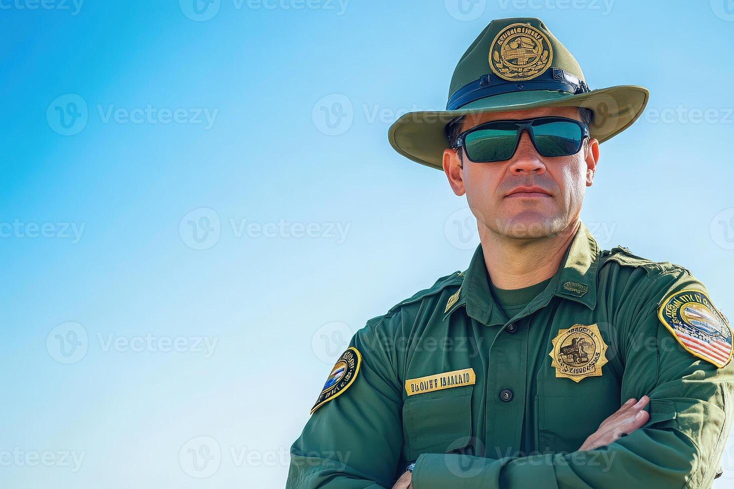 A border control officer in a green uniform and hat stands with arms crossed, wearing sunglasses. The officer is positioned against a bright blue sky, showcasing professionalism during duty. photo