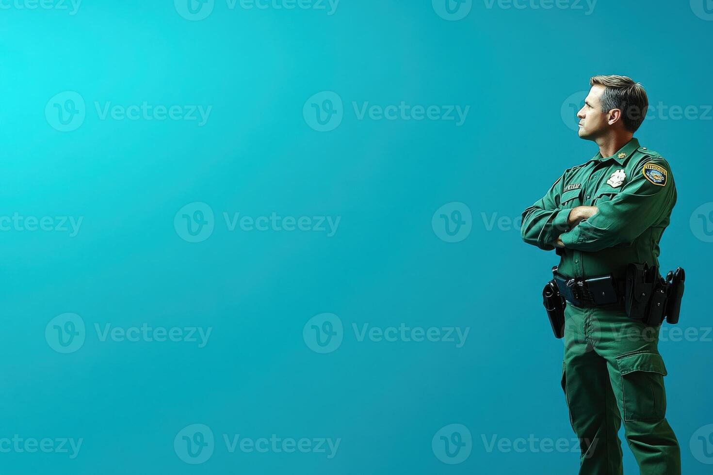 A border control officer in uniform stands with arms crossed, looking thoughtfully off to the side against a clean, solid-colored background, embodying authority and readiness in his role. photo