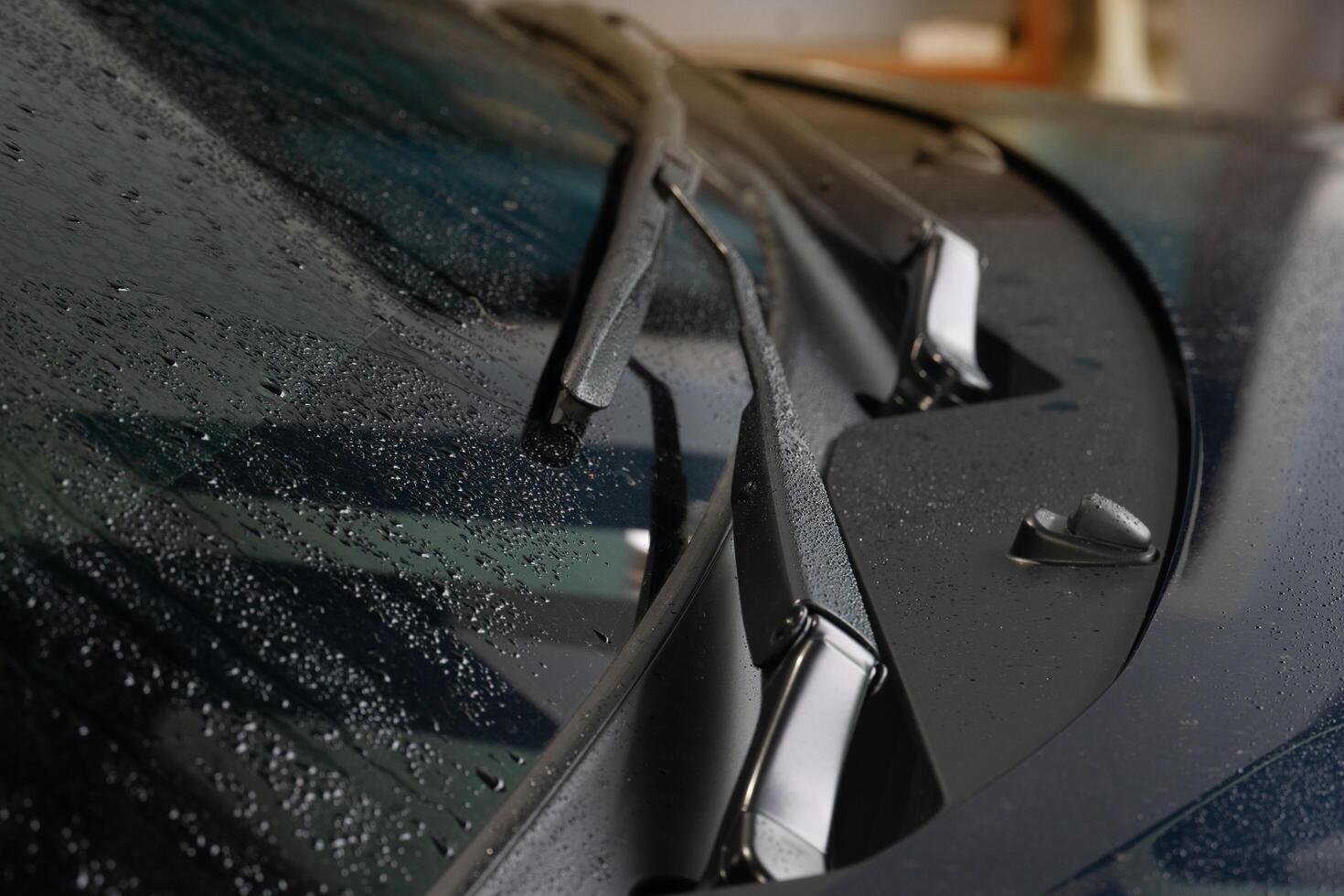 Close-up of a car windshield wiper. Wiper blade is black and appears to be made of rubber. Wiper arm is also black and has a metal spring attached to it. Background is blurred, of the hood of the car. photo
