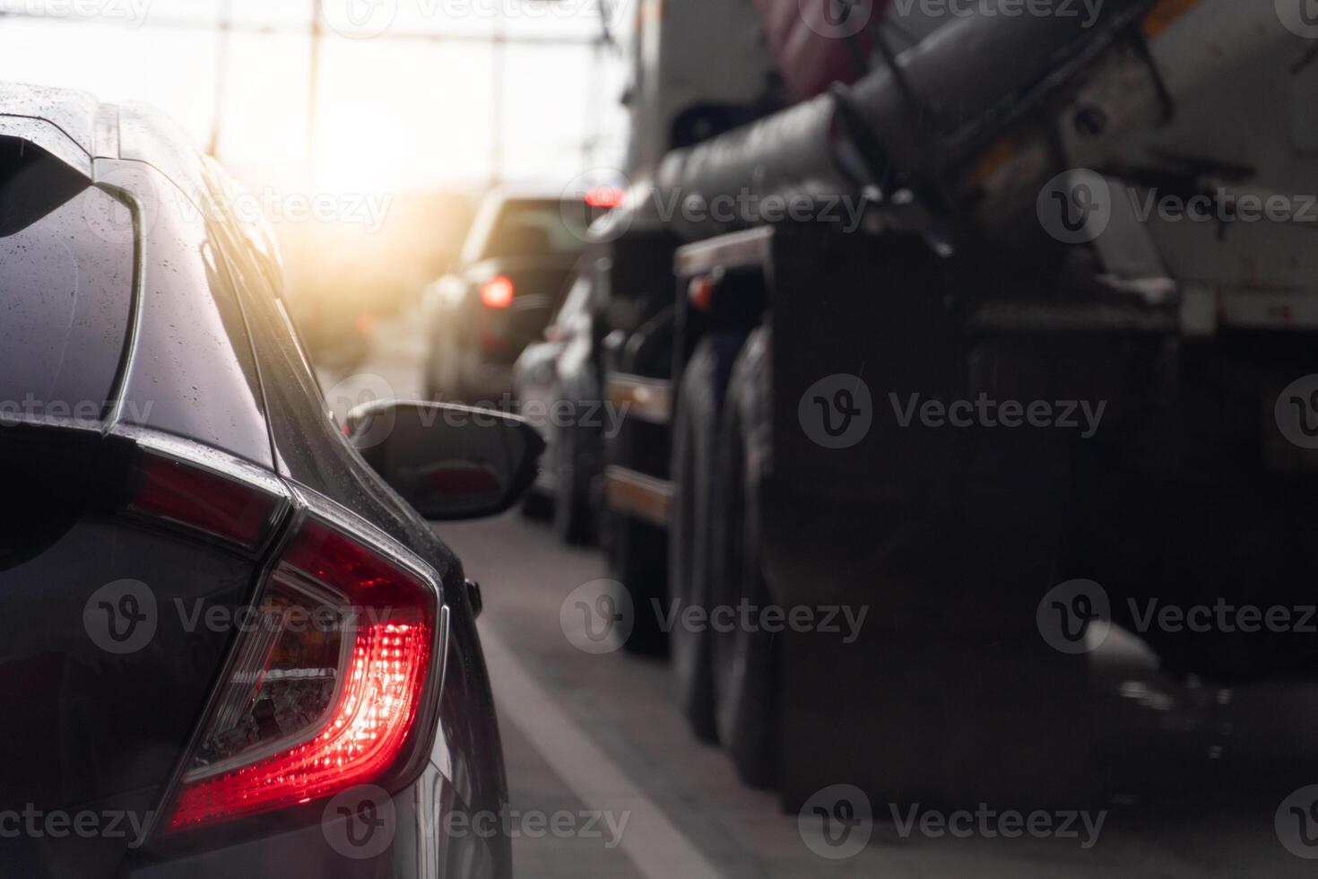 Traffic jam with several vehicles lined up on a road. Close up car with its taillights illuminated. Front of the car semi-trailer truck is partially visible. Sun is setting in the background. photo