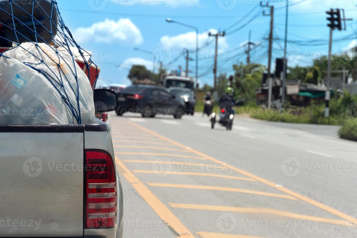 Busy on asphalt road with several vehicles in motion. Pickup truck with a load of cargo visible in the foreground. Background shows a clear blue sky with white clouds and utility poles. photo