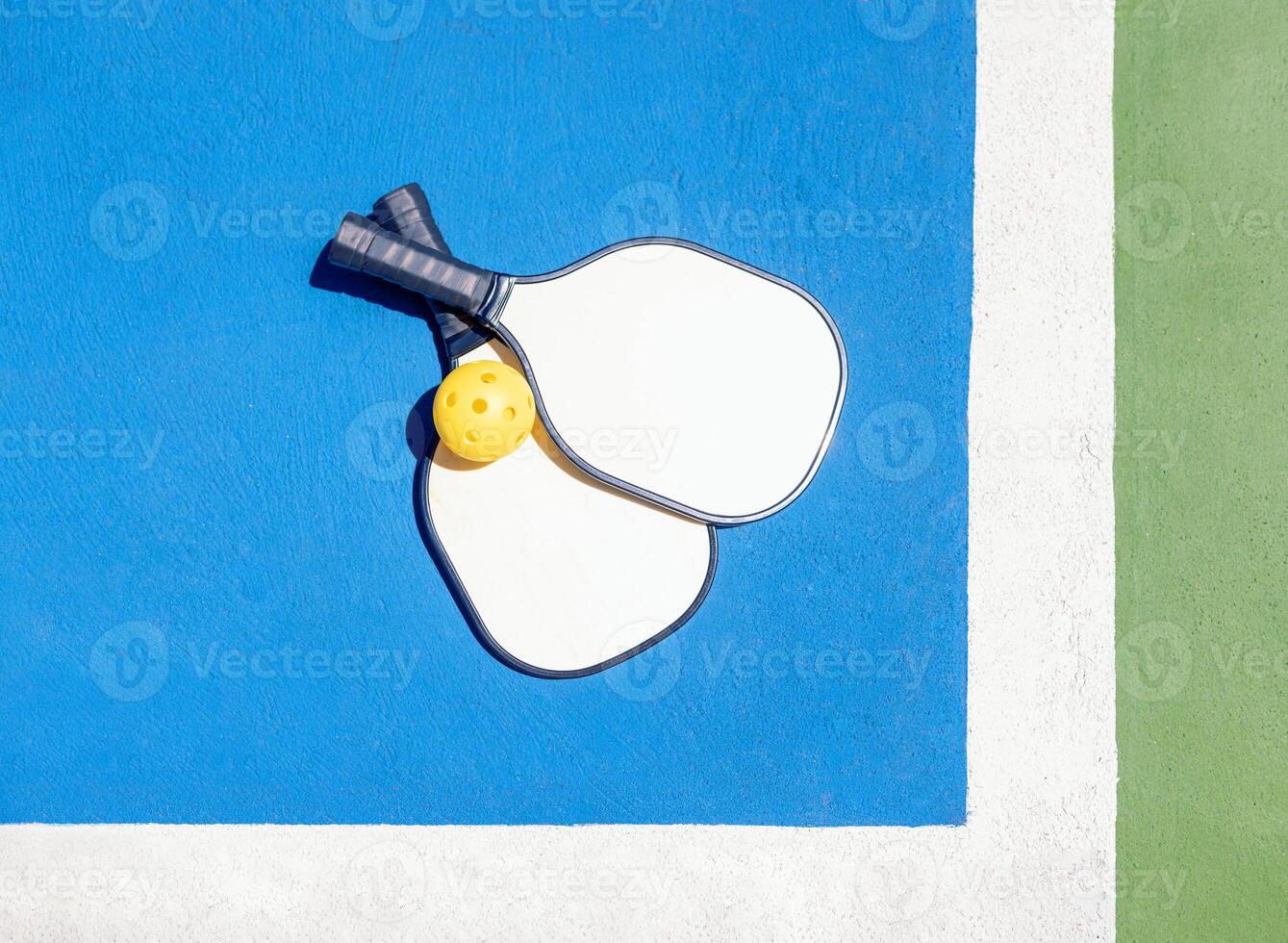 Two pickleball paddles and a yellow ball rest on a blue and green outdoor court. photo