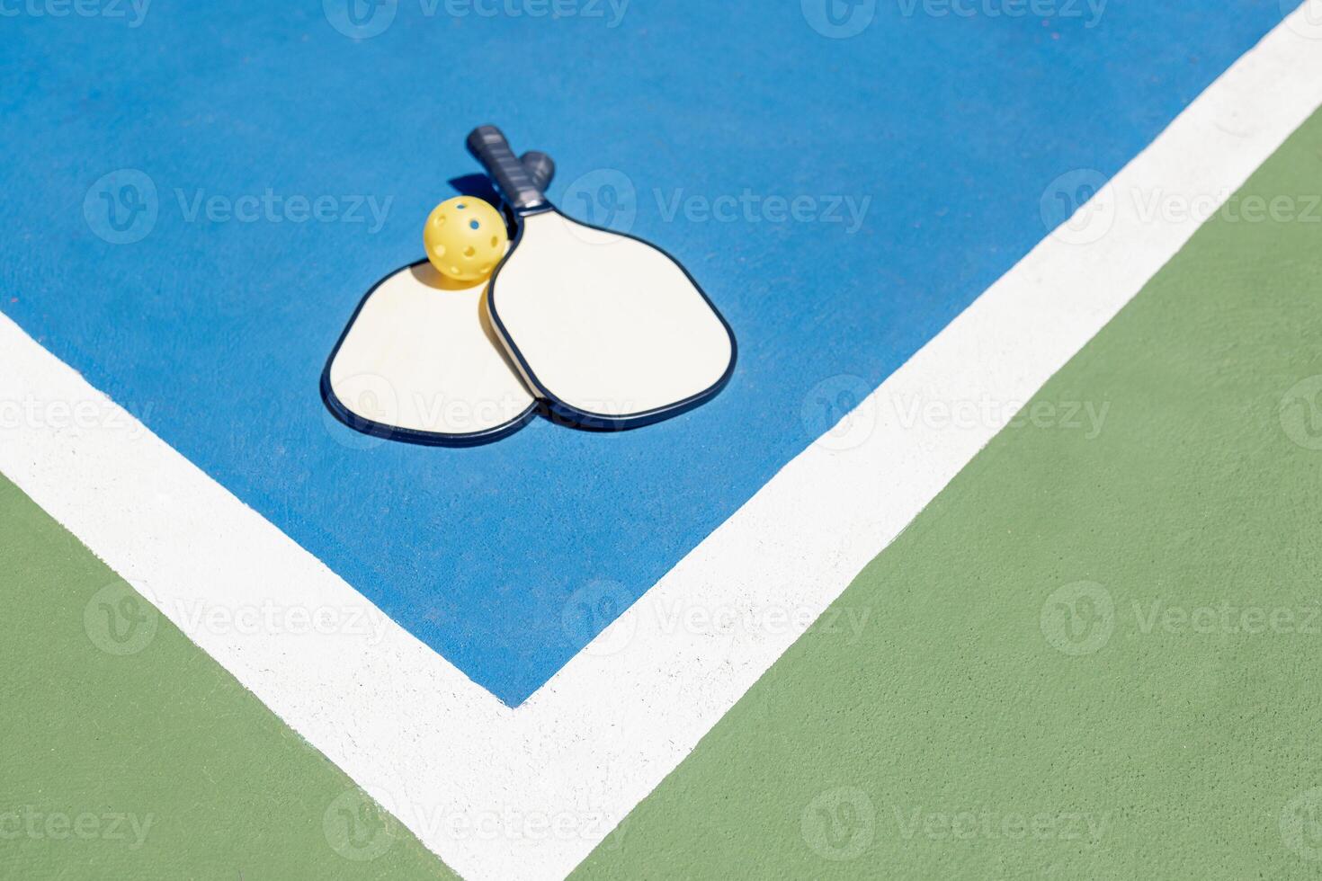 Two pickleball paddles and a yellow ball rest on a blue and green outdoor court. Contrasting colors of the playground and an energetic sports game. photo