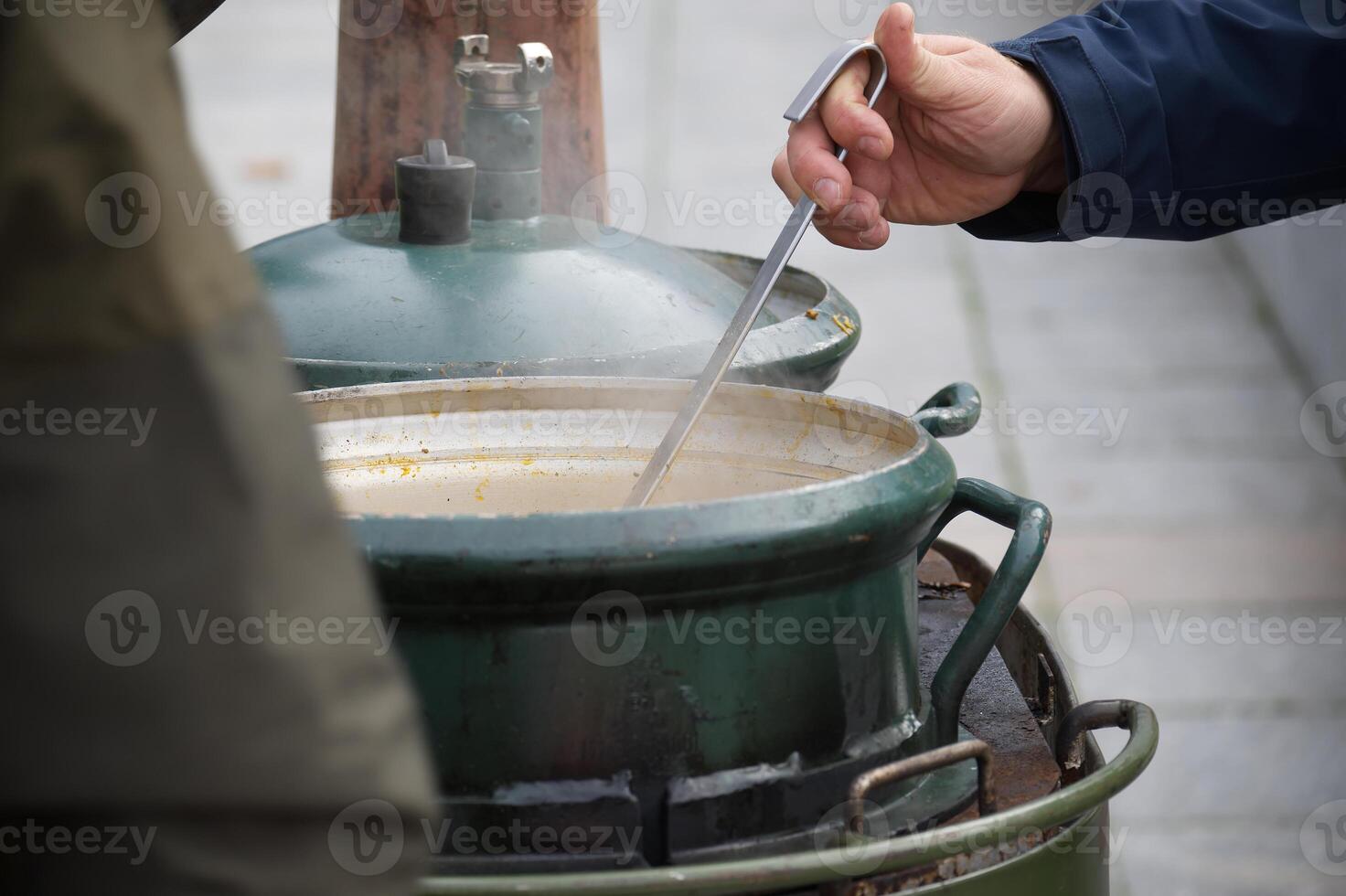 Large green metal pot with open lid and ladle serving soup outdoors on a rustic cooking setup photo