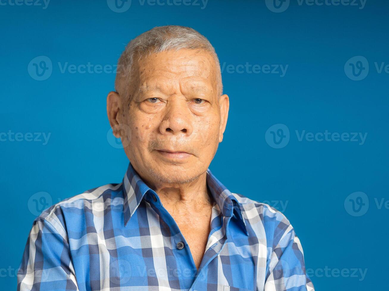 An elderly Asian man in a blue shirt looking at the camera with a smile while standing on a blue background photo