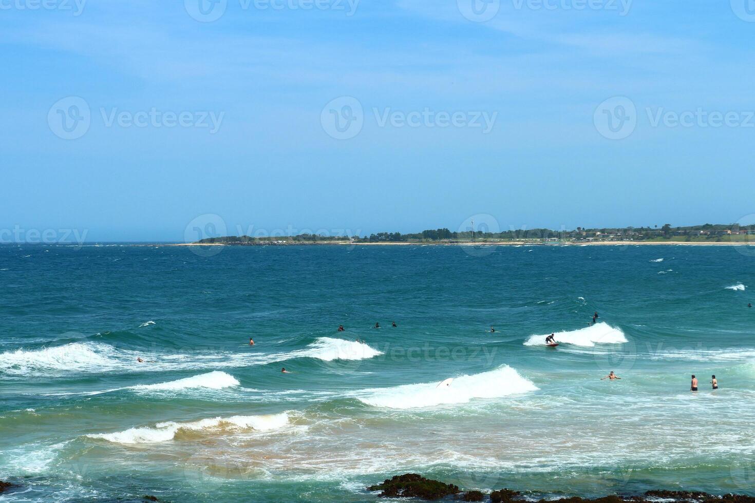 Swimmers Enjoying The Waves Of The Blue Ocean Near A Rocky Shore On A Clear Sunny Day photo