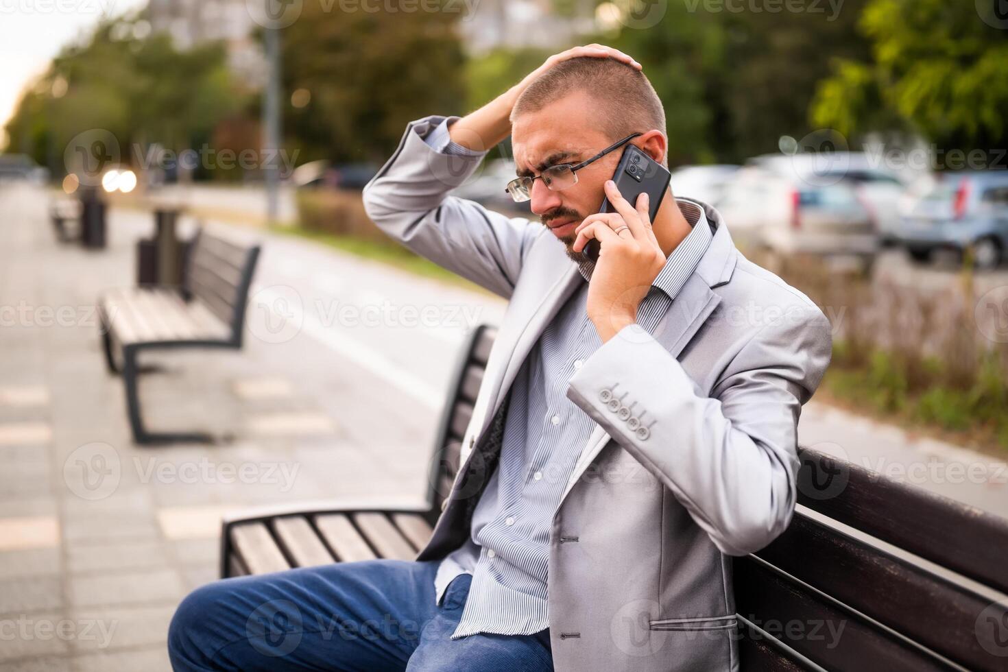 Portrait of worried businessman who is talking on phone. Man sitting on bench in the city and talking on phone. photo