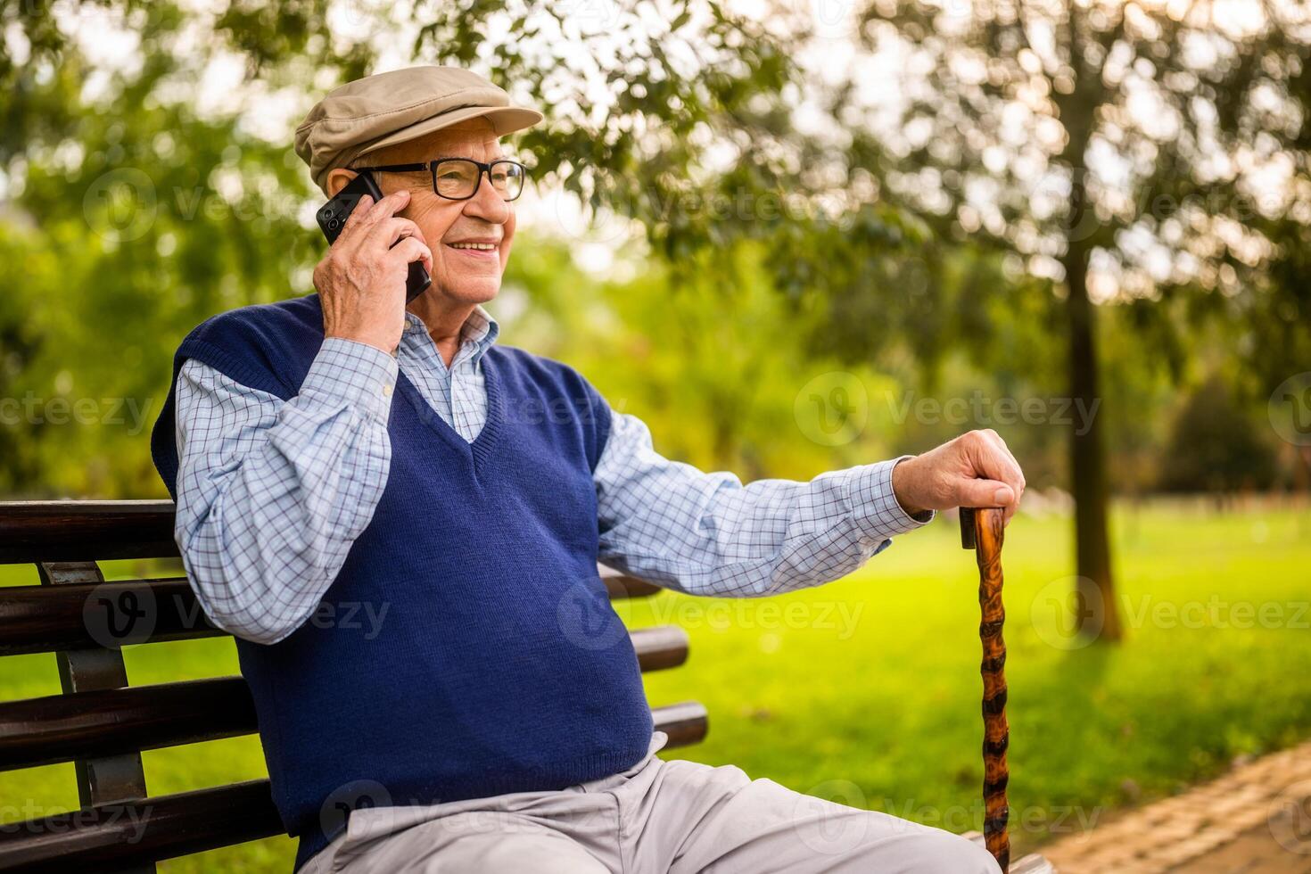 Portrait of happy senior man in park. He is sitting on bench and talking on phone. photo