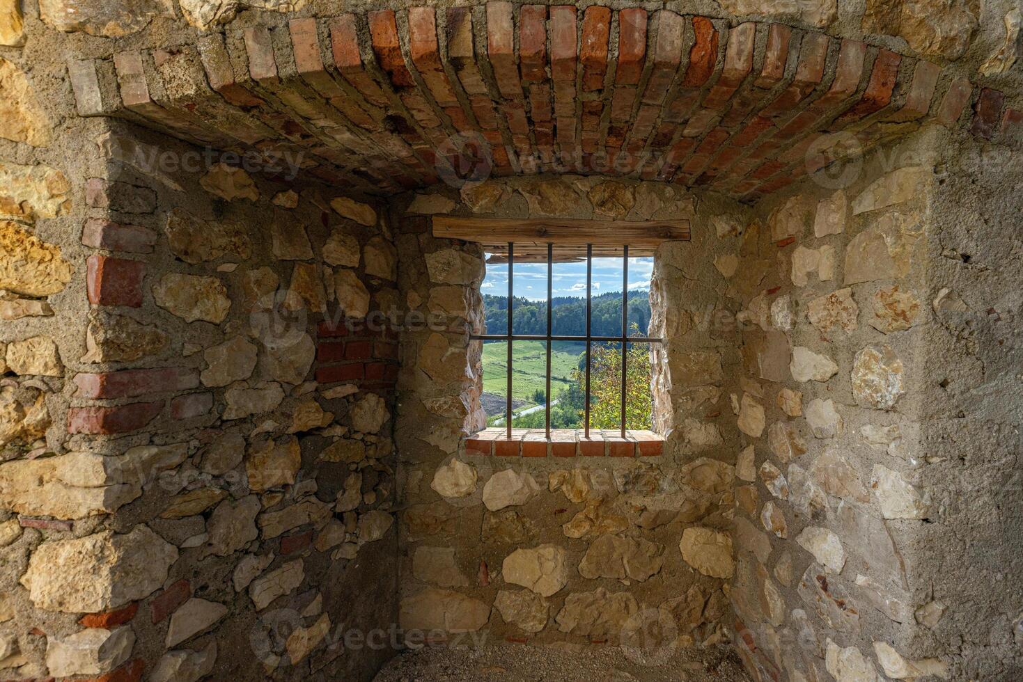 A barred window in a stone wall frames a peaceful view of green hills, fields, and distant forests photo