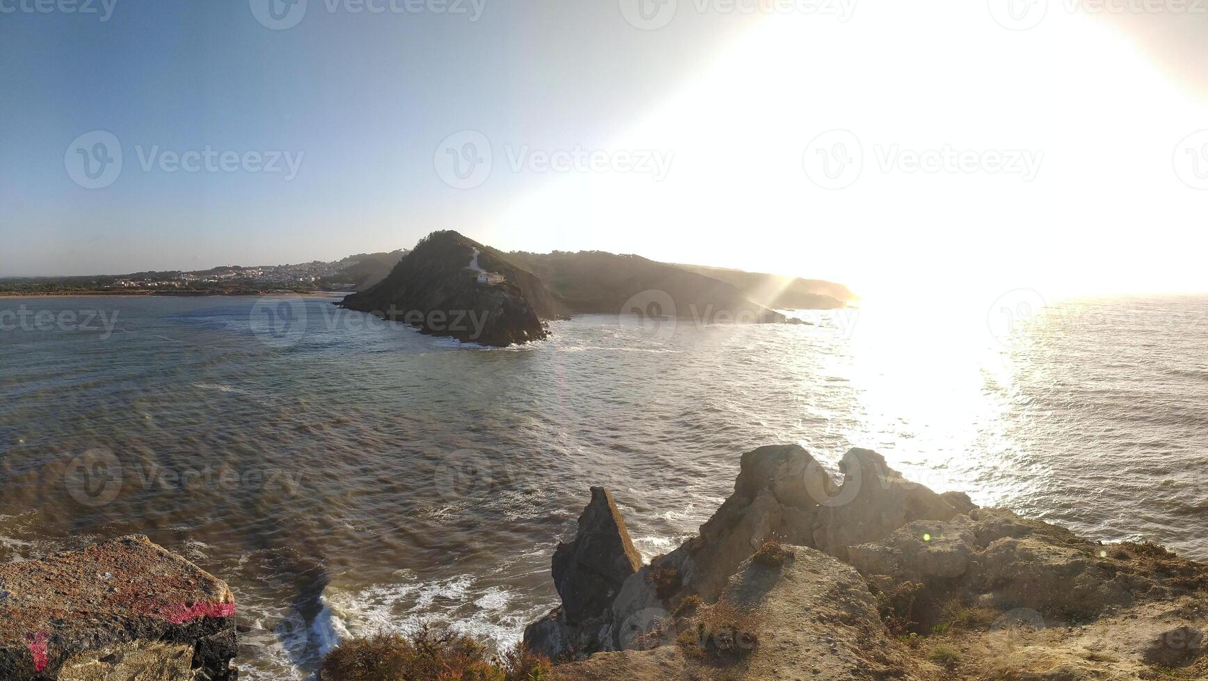 Coastal lighthouse stands on rocky terrain, with the sun setting over the ocean in a serene coastal landscape photo