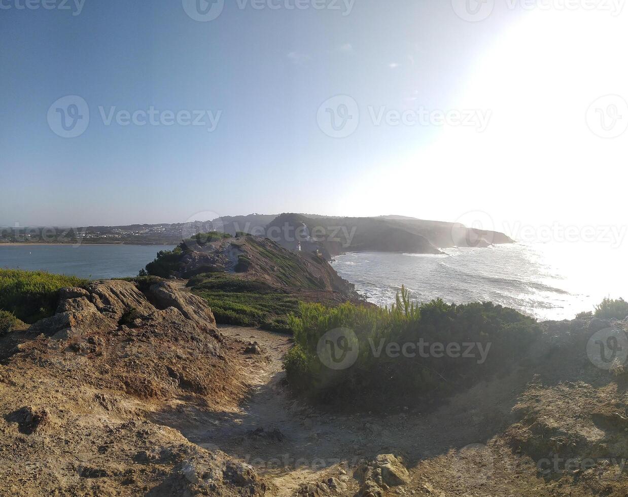 Coastal lighthouse stands on rocky terrain, with the sun setting over the ocean in a serene coastal landscape photo