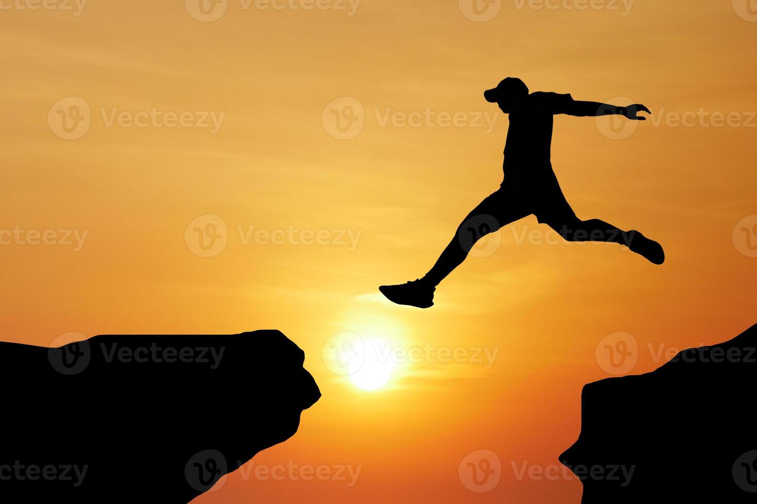 Silhouette of a Person Leaping Across a Gap at Sunset with a Vibrant Orange Sky in the Background photo