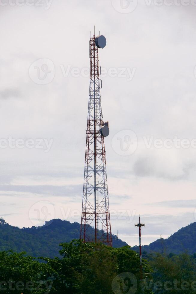 Tall Communication Tower Against a Cloudy Sky with Lush Greenery and Distant Hills in the Background photo