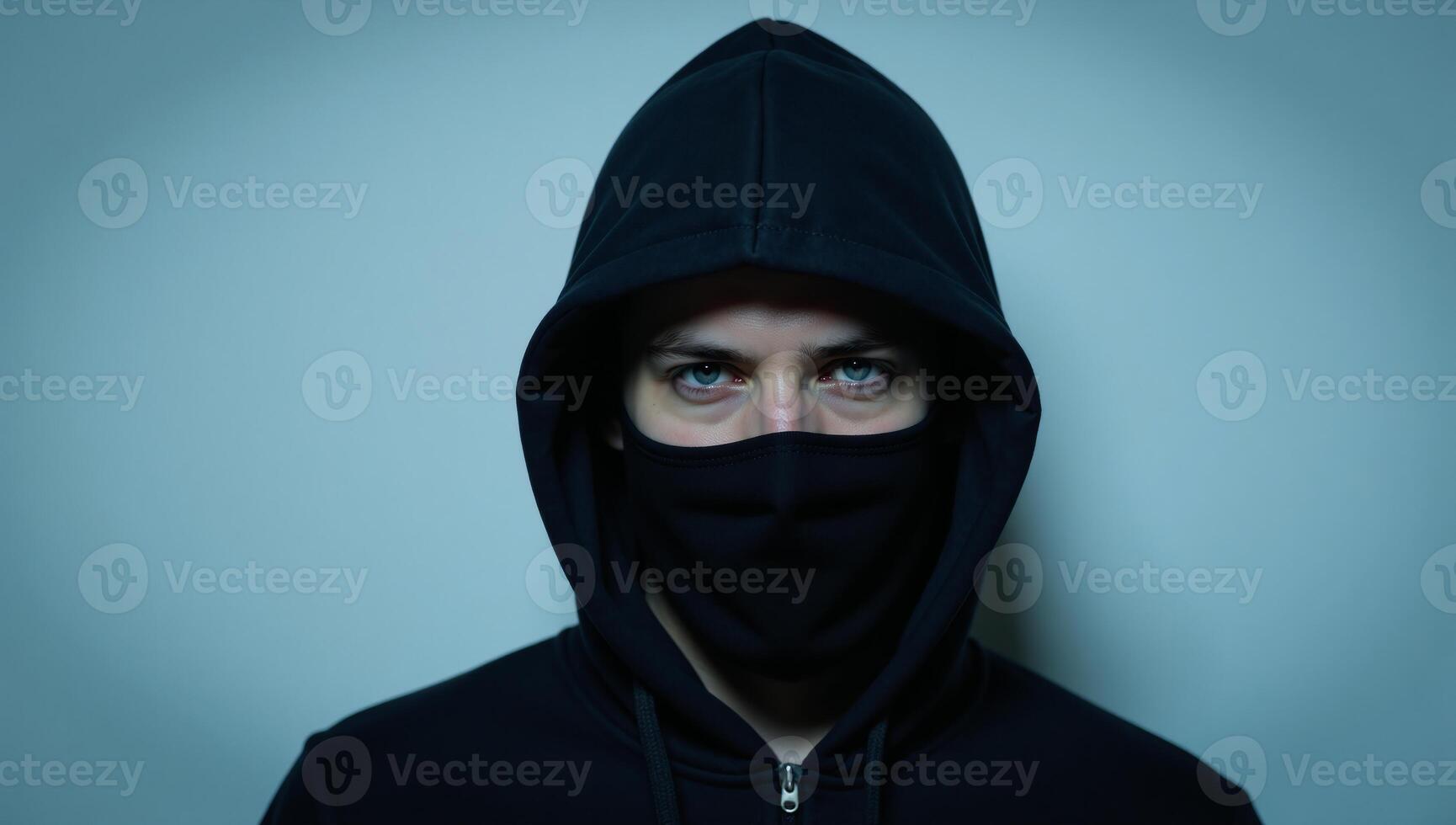 A darkhoodied masked man with piercing blue eyes looks straight into the camera photo