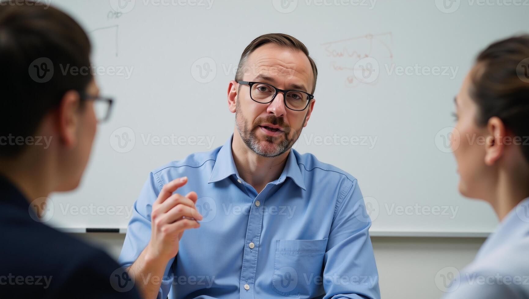 A bearded man at a desk discusses ideas with two others while standing before a whiteboard photo