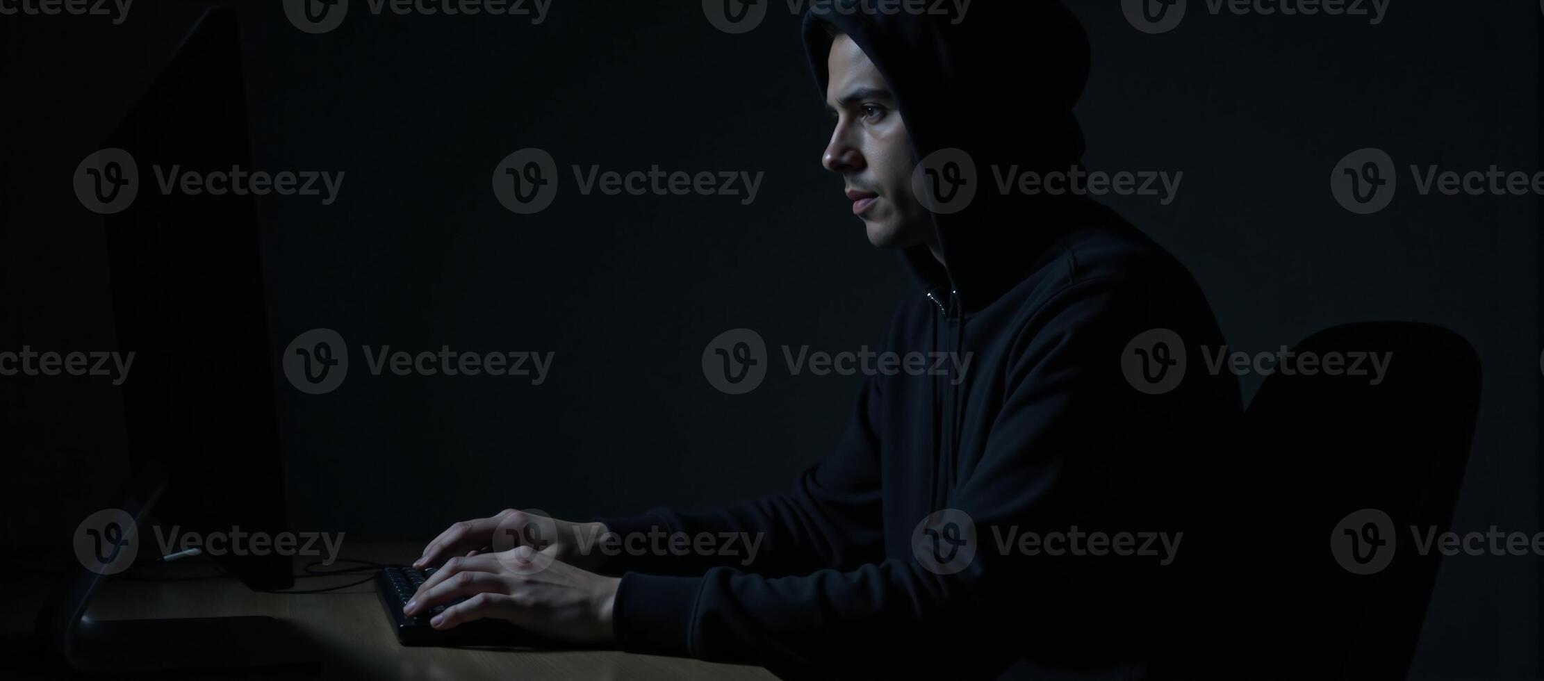 An anonymous figure in a hoodie uses a computer within a dimly lit room evoking a sense of cybercrime intrigue photo