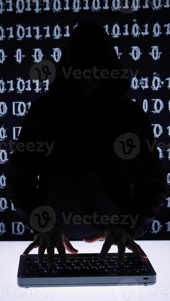 A hooded criminal silhouetted while hacking on binary code background cybercrime concept photo
