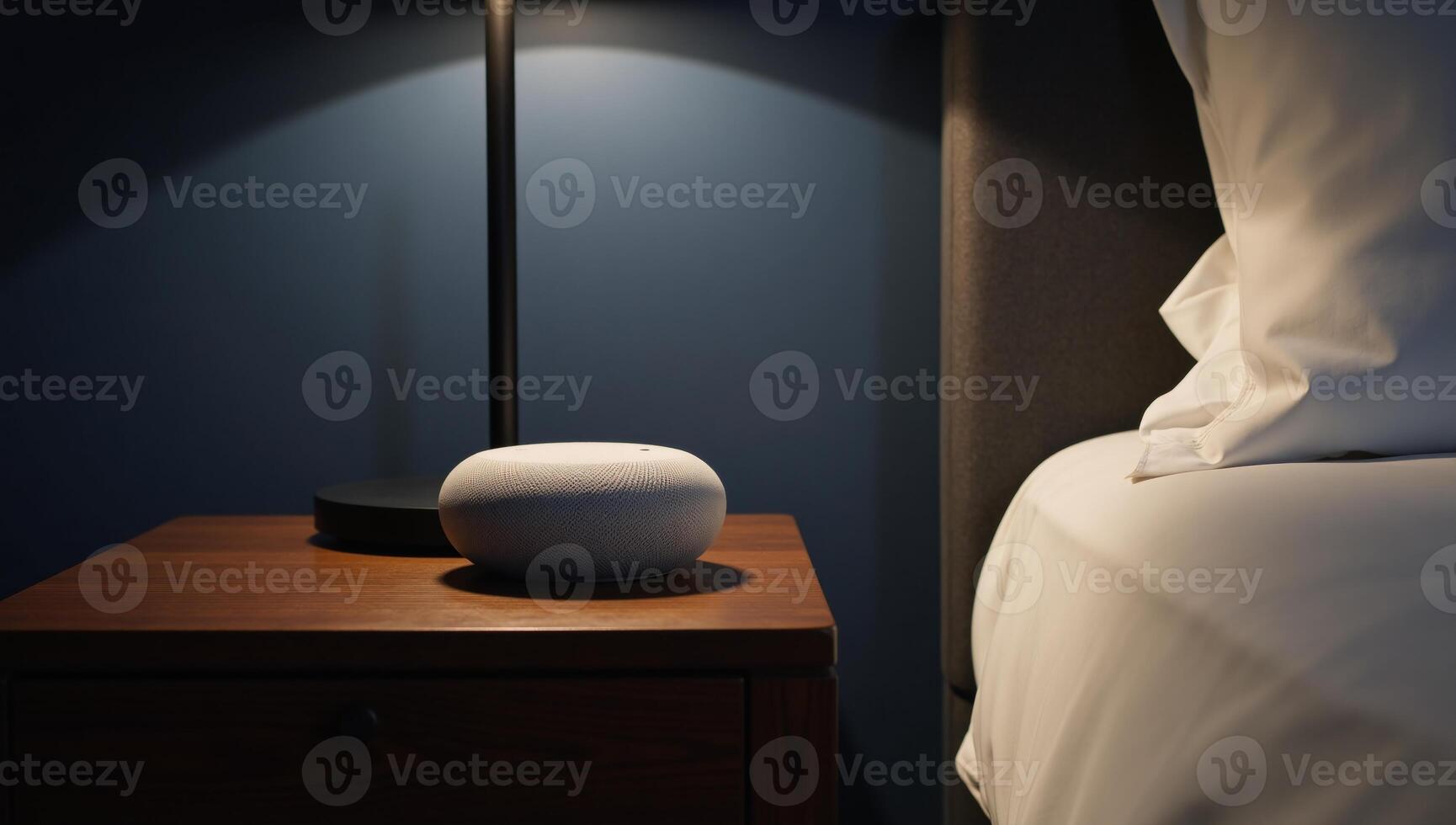 Sleek greywhite smart speaker sits atop lit nightstand in dark blue bedroom photo