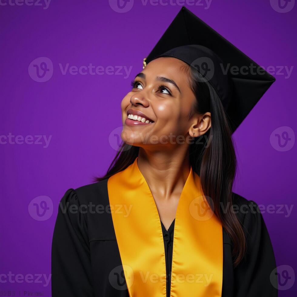 A smiling thoughtful young AfricanAmerican woman is depicted in a graduation cap and gown standing before a vibrant purple backdrop photo
