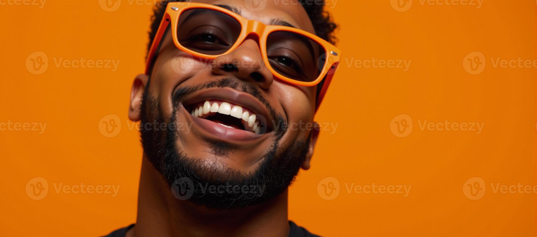 A bearded black man with orange glasses grins at the camera against an energetic orange backdrop photo