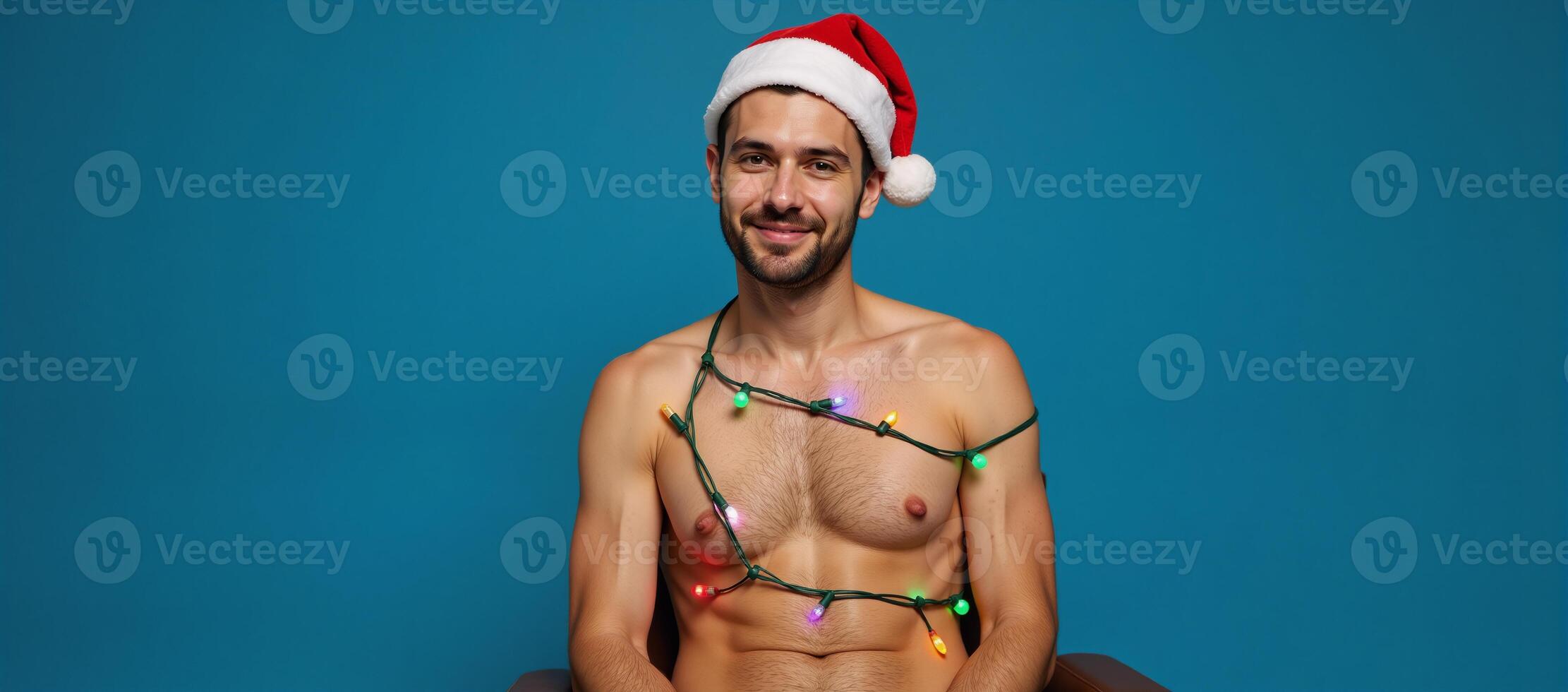 A portrait featuring a gay man seated against a blue backdrop adorned with Christmas lights and wearing a Santa Claus hat for Christmas Eve photo