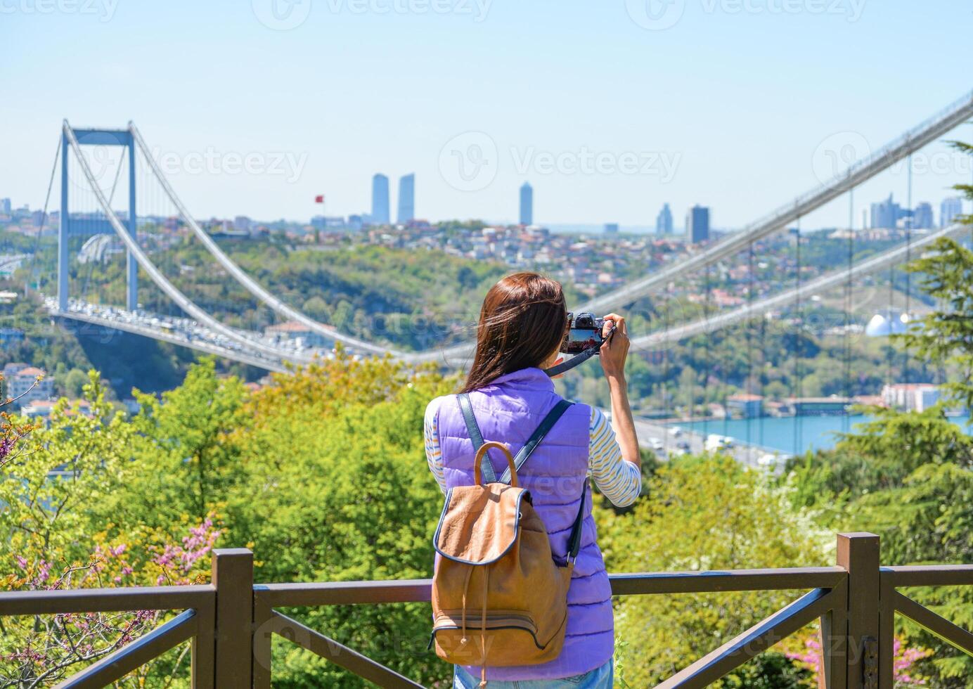 A young woman tourist with a backpack photographs the cityscape with a panoramic view of the Fatih Sultan Mehmet Bridge. Back view. Otagtepe Park photo