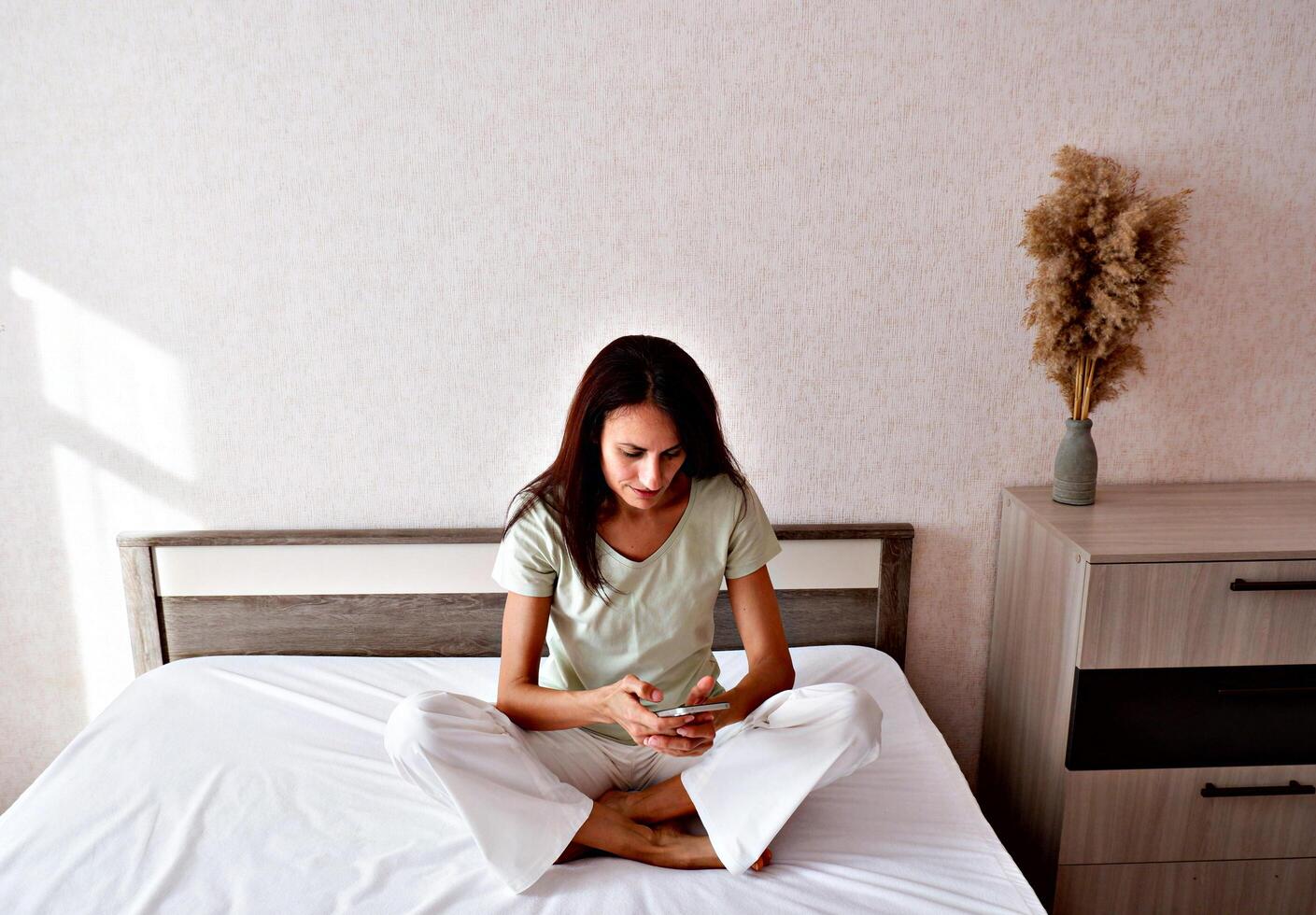 Brunette woman sitting in white bed using smartphone, having call, smiling young woman relaxing in bedroom, waking up in the morning and sending text messages on one device, technology concept photo