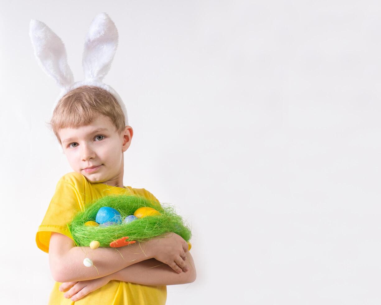 Happy child smiling in a yellow t-shirt in rabbit ears and with colorful eggs in a basket on a white background. Happy Easter concept with place for text. photo