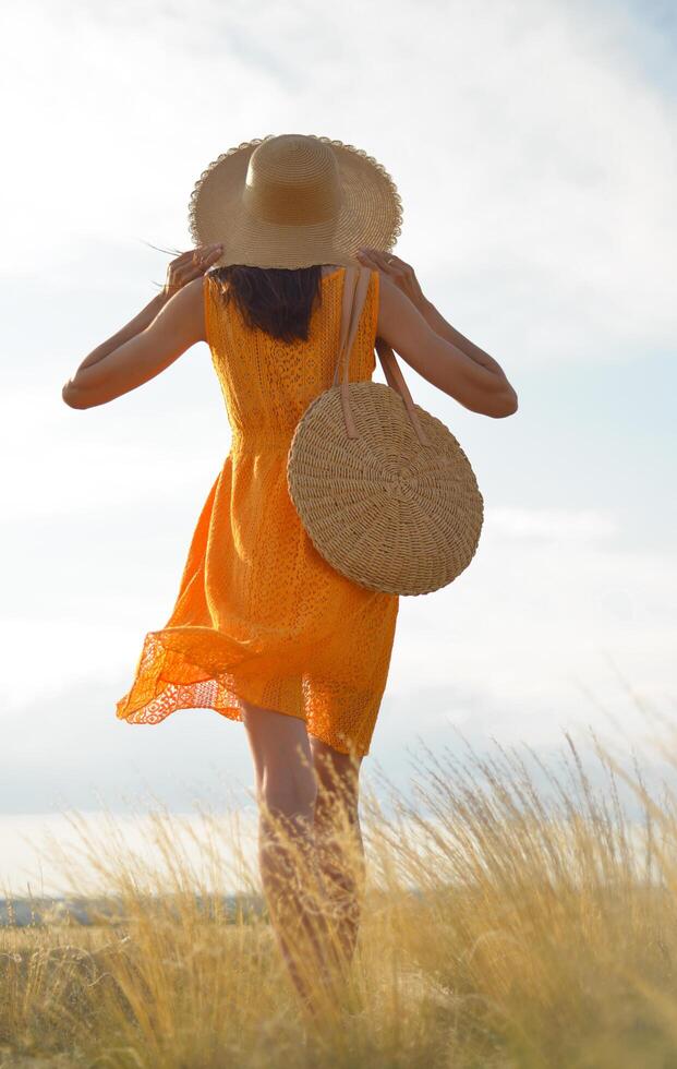 of a beautiful girl dressed in a casual orange dress with a straw hat and a straw bag in her hands in a field in the sunlight. Blows long hair. Autumn. Shine the sun, sunshine. backlit Warm tinted photo