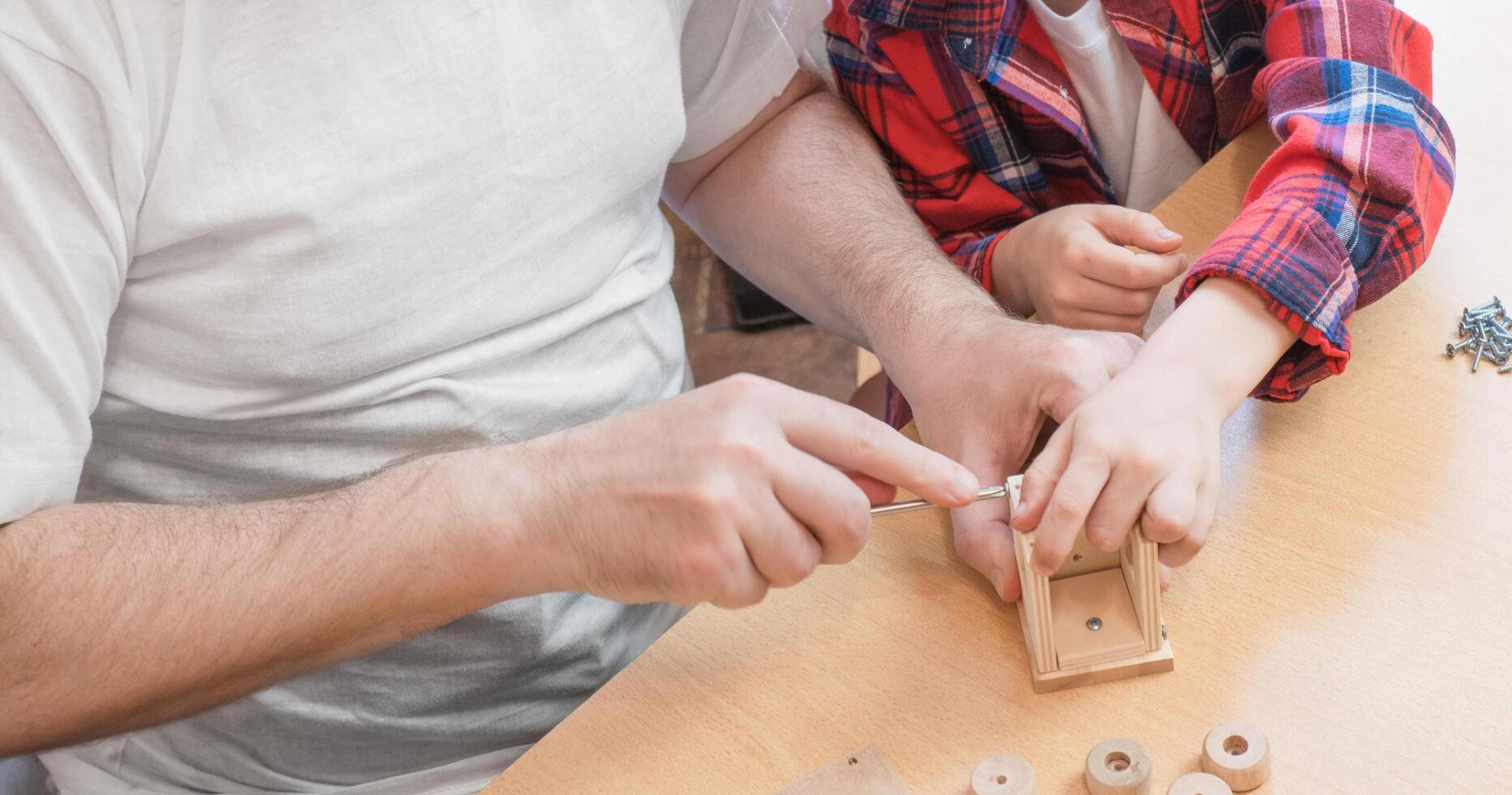Happy father's day and childhood concept. Close-up of a father and a boy son in glasses work with hand tools, using a screwdriver, assembling a wooden house constructor at the table. photo