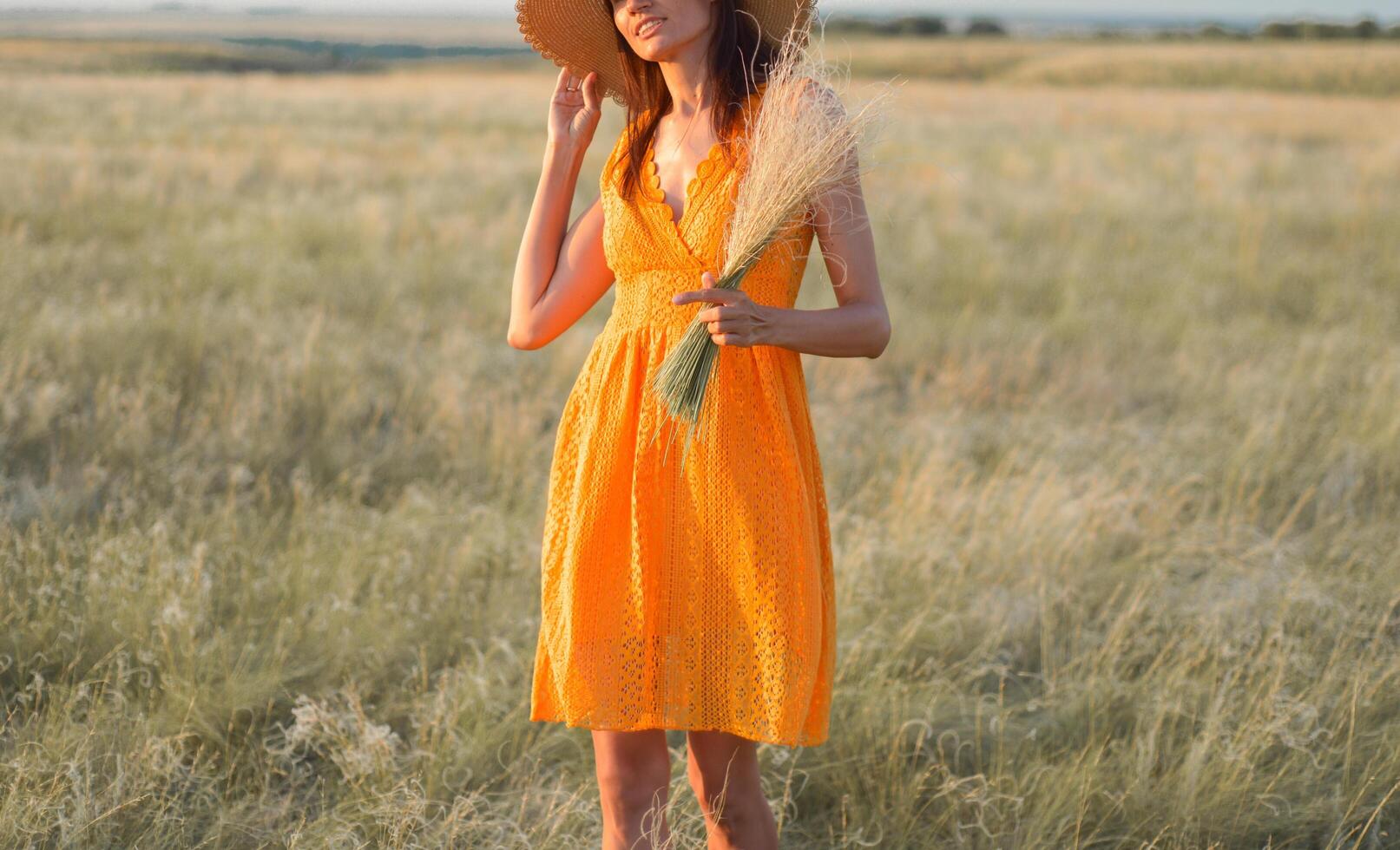 Young woman in an orange dress and a straw hat standing on a field in the rays of the setting sun photo