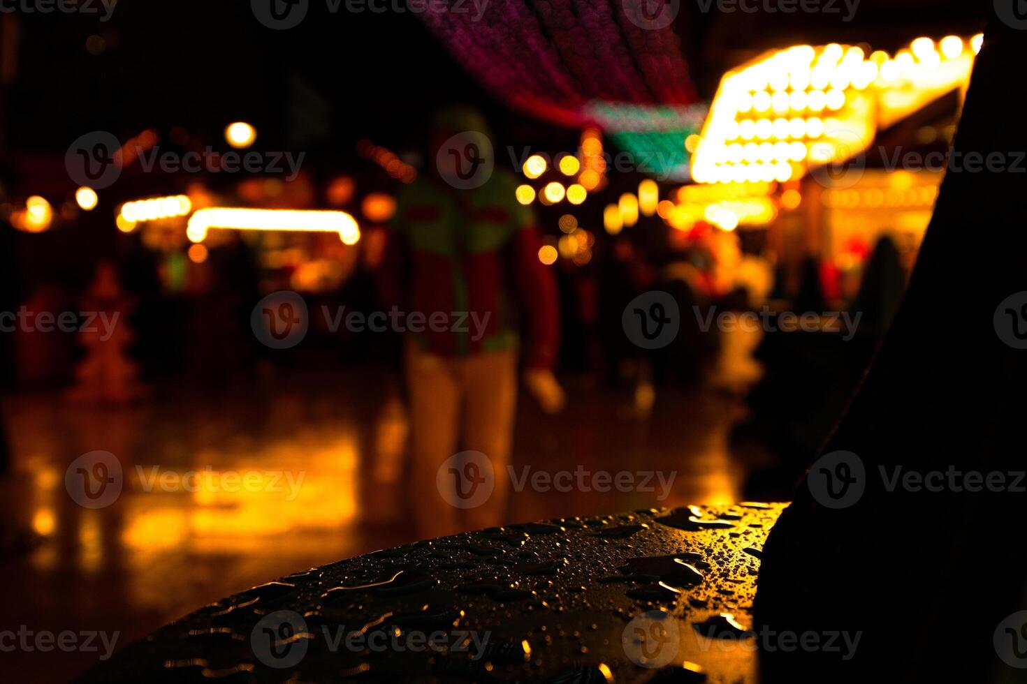Rainy night at Christmas matket. Raindrops on a the table surface and blurry festive lights in the blurred background. Persons silhouette and distant people walking down the illuminated street photo