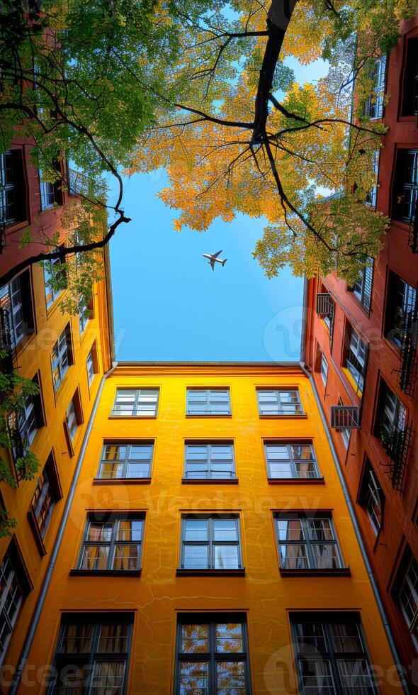 Vibrant courtyard with trees and planes A vibrant courtyard framed by autumn leaves and colorful buildings under a clear blue sky, showcasing an airplane flying overhead. photo