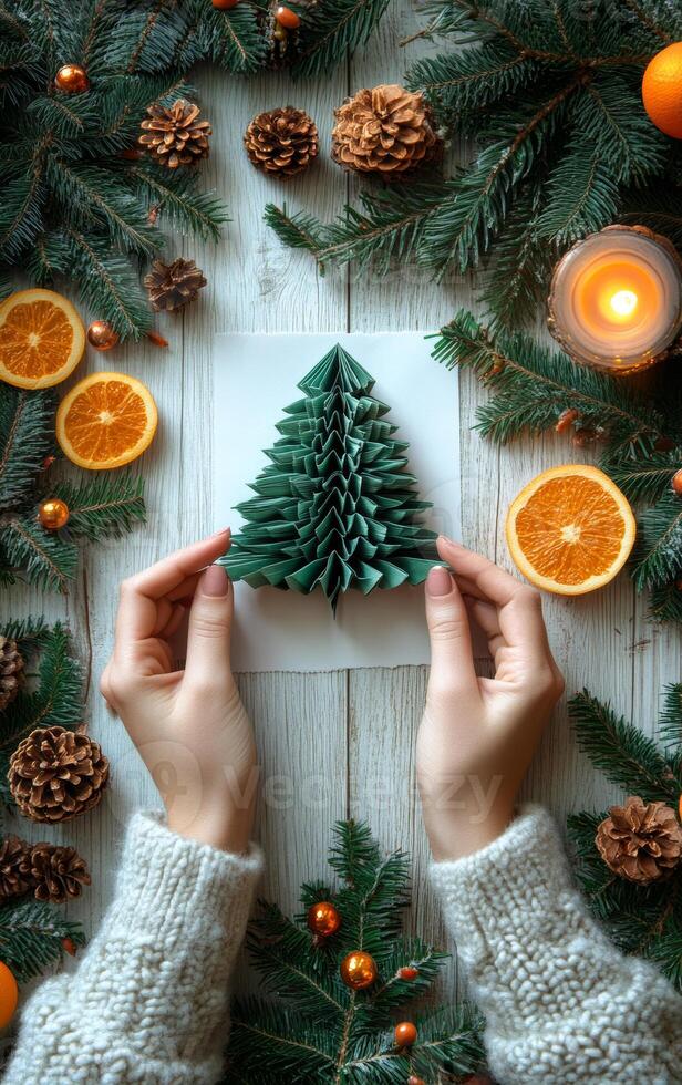 Making a decorative paper Christmas tree. A person is creating a green paper Christmas tree surrounded by orange slices, pine cones, and candles on a wooden table. photo
