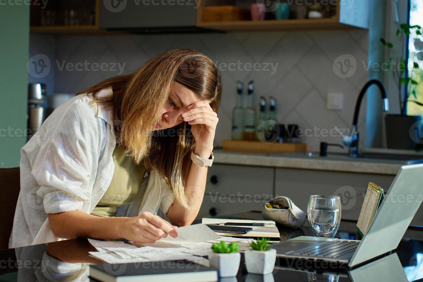 Portrait of stressed woman sitting in kitchen with laptop and stack of paper invoice. Female planning home budget, looking at finance bill and calculating expenses photo