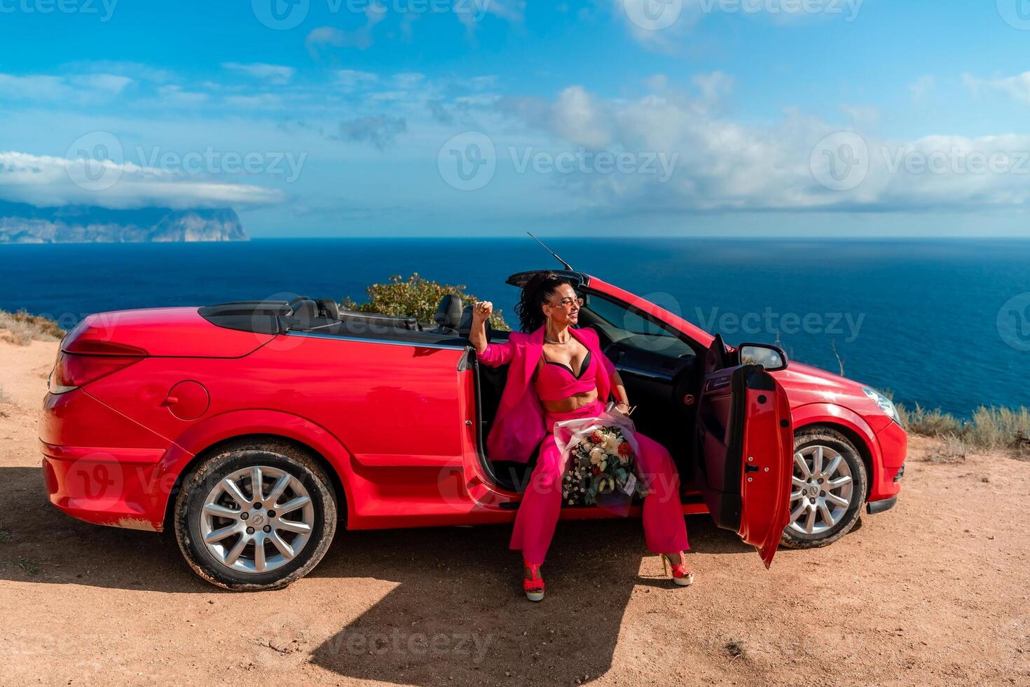 A woman is sitting in the back of a red convertible car. The car is parked on a dirt road near the ocean. The woman is wearing a pink suit and red shoes. Scene is relaxed and carefree. photo